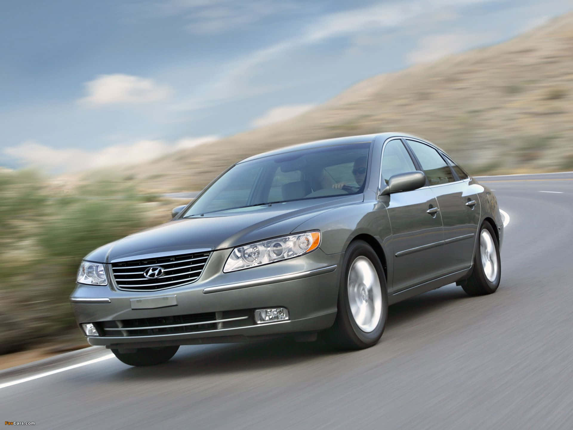 Hyundai Azera 桌布 壁紙 — 一輛時尚的銀色現代轎車在蜿蜒的道路上行駛，背景是模糊的山丘和天空。