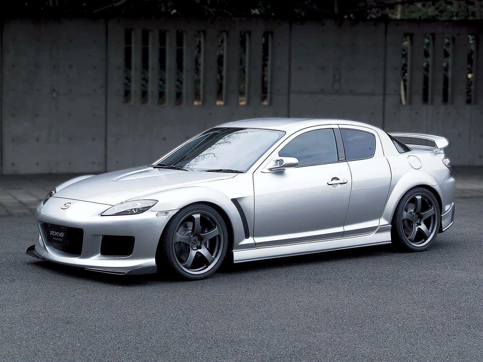 Mazda Rx-8 桌布 壁紙 — 一輛時尚的銀色萬事得RX-8跑車，配備黑色輪圈，停在瀝青路面上，背景是帶有垂直縫隙的混凝土牆。