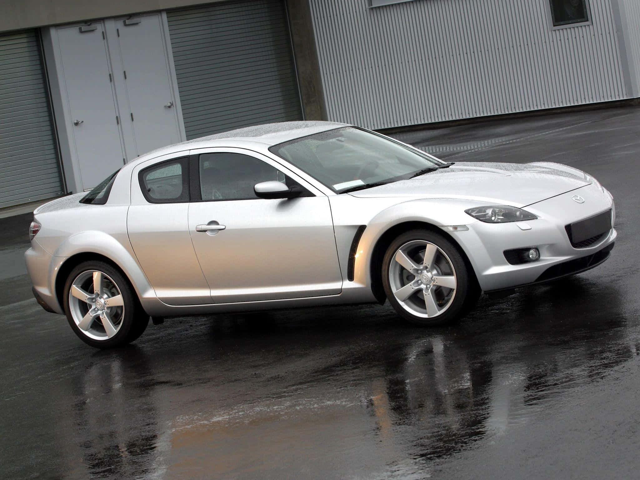 Mazda Rx-8 桌布 壁紙 — 一輛時尚的銀色萬事得RX-8轎跑車在濕瀝青路面上閃閃發光，其曲線倒映在灰色工業建築背景下。