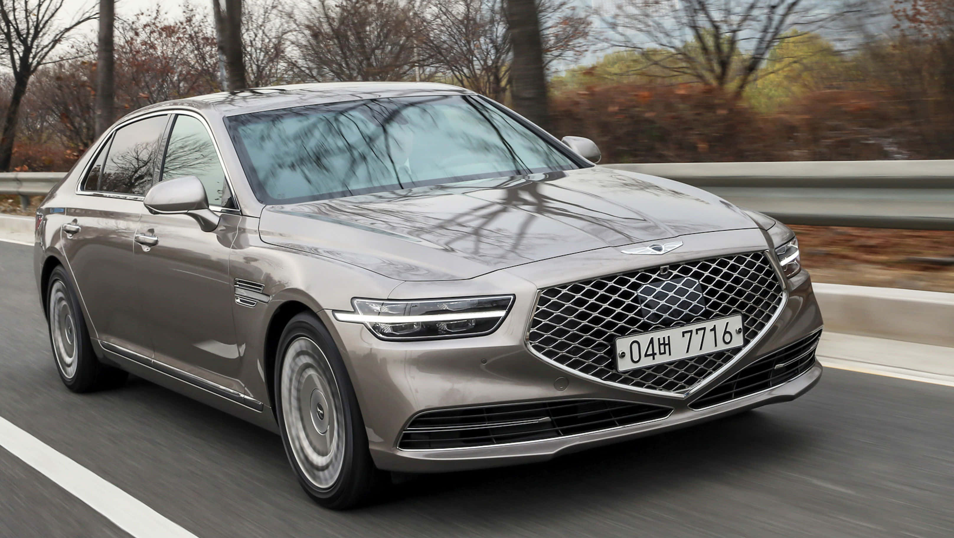 Genesis G90 桌布 壁紙 — 一輛時尚的銀色豪華轎車在兩旁種滿樹木的道路上行駛，秋天的樹葉呈現動態模糊效果。