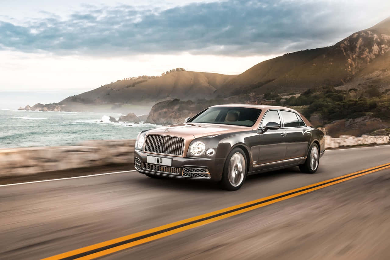 Bentley Mulsanne 桌布 壁紙 — 一輛時尚的銀色賓利慕尚沿著海岸公路行駛，兩旁是崎嶇的懸崖和海洋波浪，天空烏雲密佈，氣氛戲劇化。