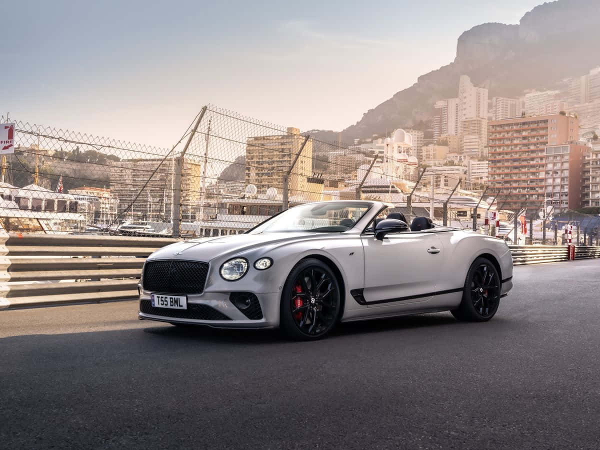 Bentley Continental Gt 桌布 壁紙 — 一輛時尚的銀色賓利開篷車，帶有黑色飾邊和紅色制動卡鉗，停在俯瞰摩納哥海岸城市景觀的賽道上，正值黃金時刻。