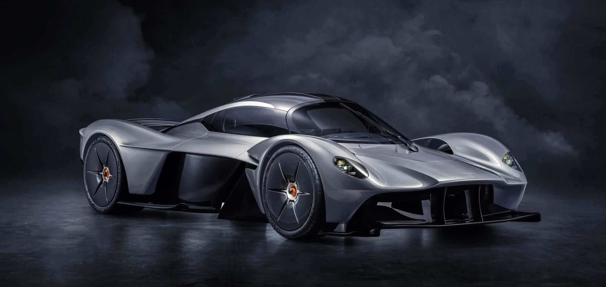 Aston Martin Valkyrie 桌布 壁紙 — 一輛時尚的銀色超級跑車，配備發光的車頭燈，在反射性地板上，以黑暗煙霧繚繞的背景戲劇性地矗立。