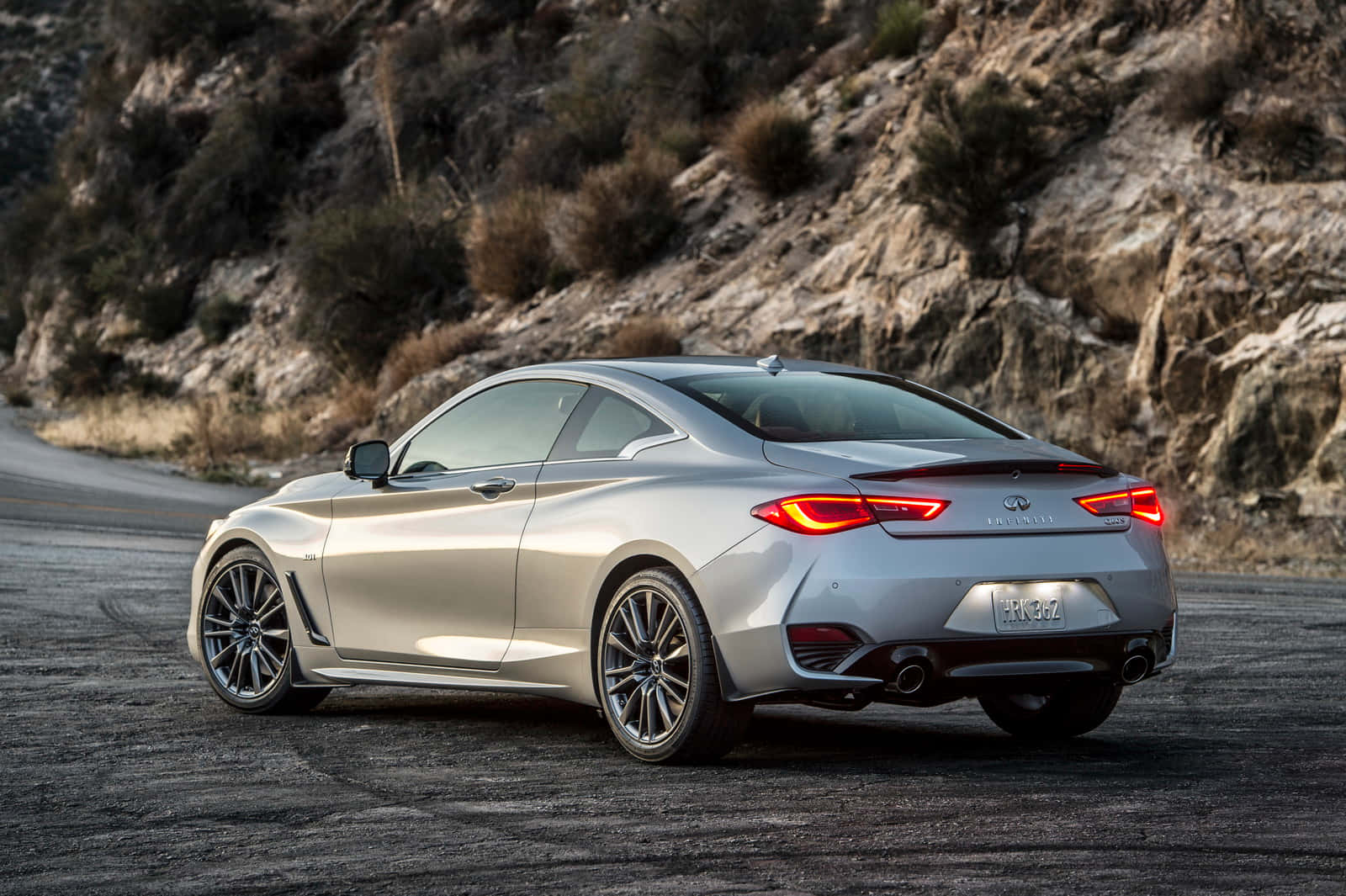 Infiniti Q60 桌布 壁紙 — 一輛時尚的銀色跑車，帶有發光的紅色尾燈，停放在蜿蜒的山路上。