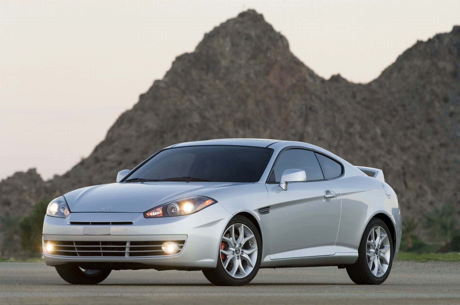 Hyundai Tiburon 桌布 壁紙 — 一輛時尚的銀色跑車停在沙漠公路上，背景是岩石山脈和日落。