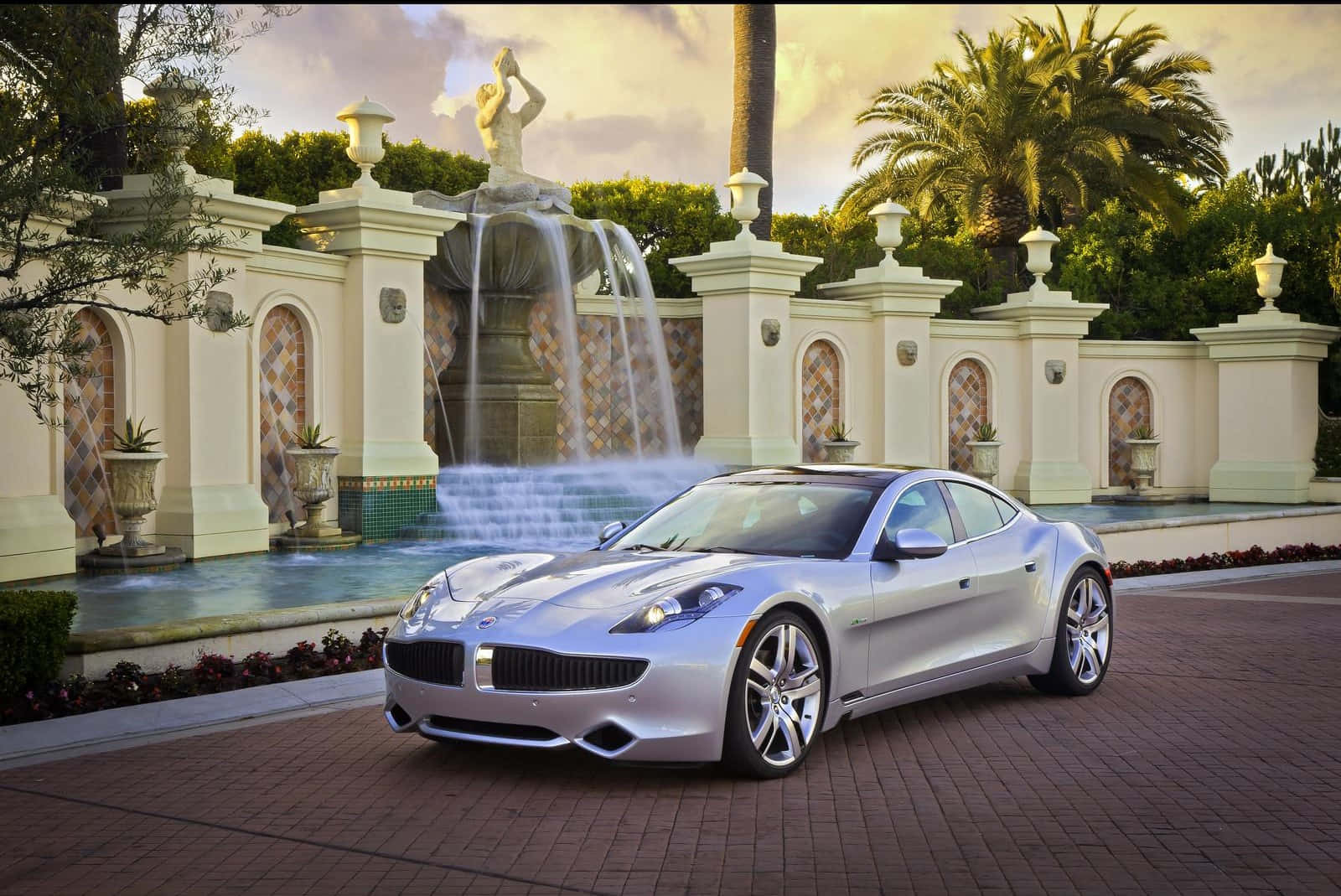 Fisker 桌布 壁紙 — 一輛時尚的銀色跑車停在磚砌車道上，前面是裝飾華麗的噴泉，有雕像和棕櫚樹，背景是金色的日落天空。