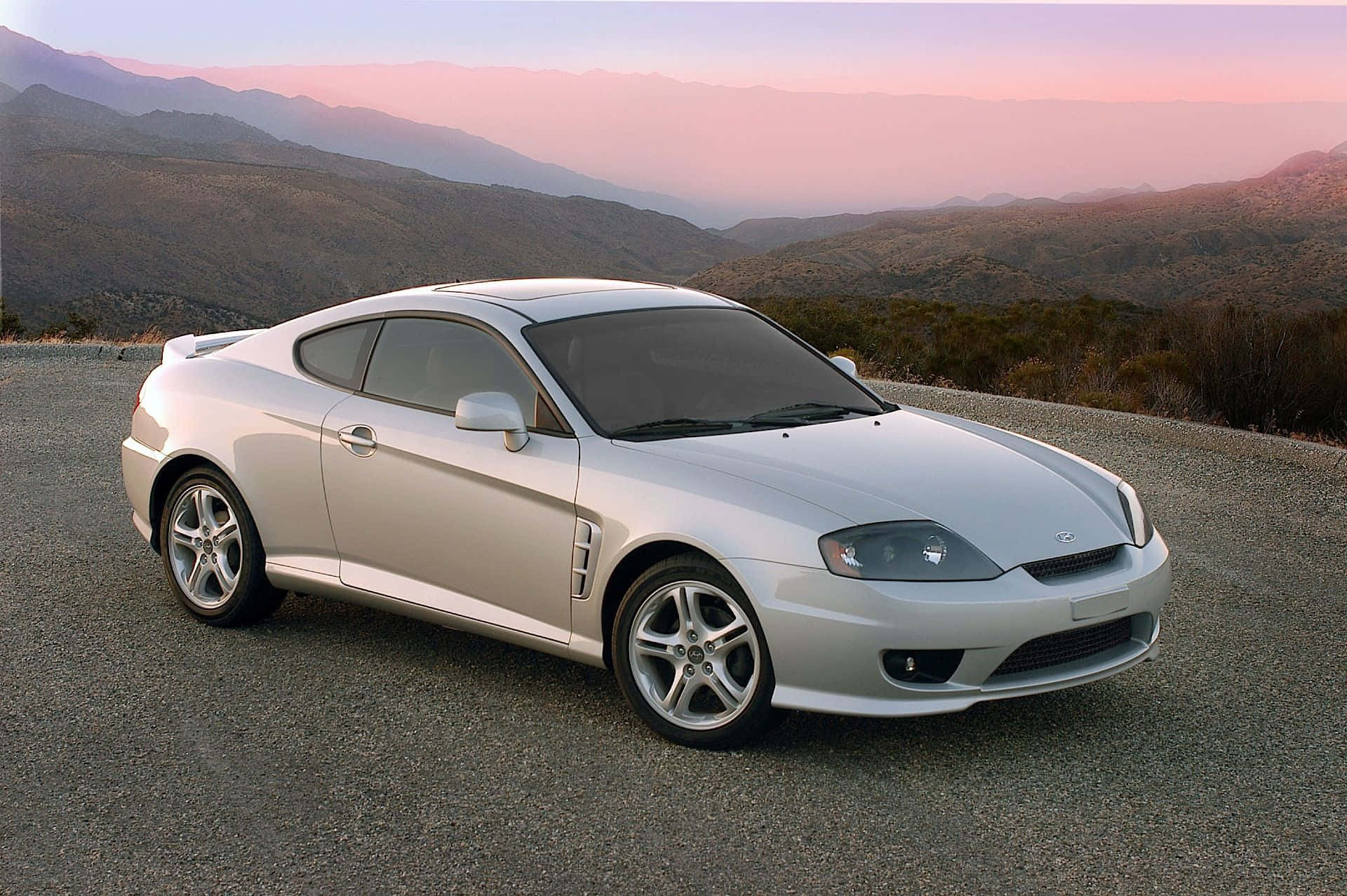 Hyundai Tiburon 桌布 壁紙 — 一輛時尚的銀色跑車停泊在蜿蜒的山路上，夕陽映照著粉紅和橙色的天空。