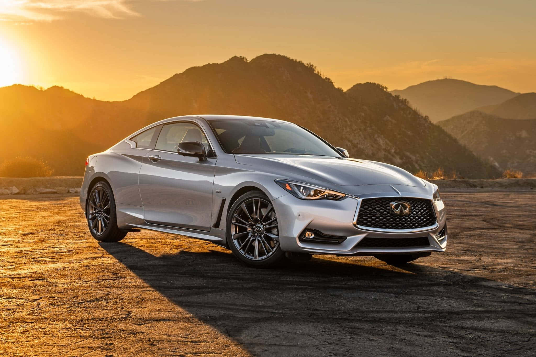 Infiniti Q60 桌布 壁紙 — 一輛時尚的銀色跑車停泊在陽光明媚的沙漠公路上，群山沐浴在金黃色的日落餘暉中。