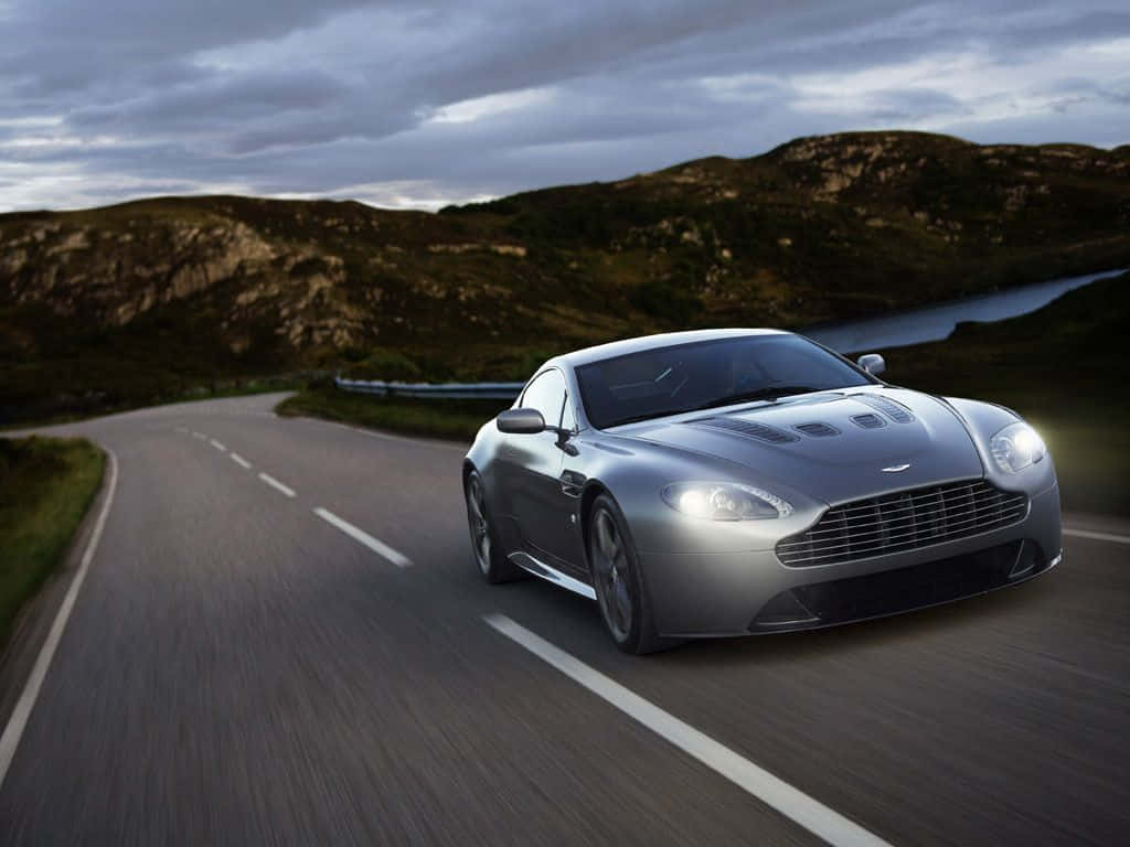 Aston Martin V12 Vantage 桌布 壁紙 — 一輛時尚的銀色跑車在多雲的黃昏天空下沿著蜿蜒的山路疾馳。