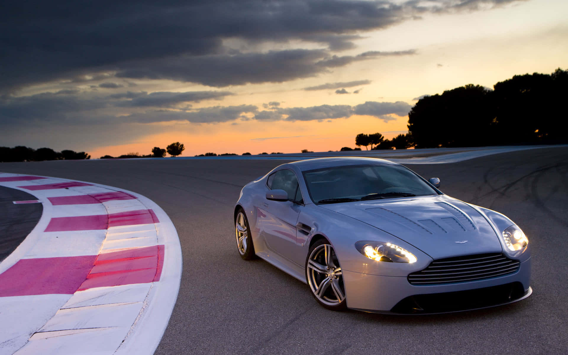 Aston Martin V12 Vantage 桌布 壁紙 — 一輛時尚的銀色跑車在日落時分，伴隨著戲劇性的雲彩和發光的車頭燈，在賽道彎道上滑行。
