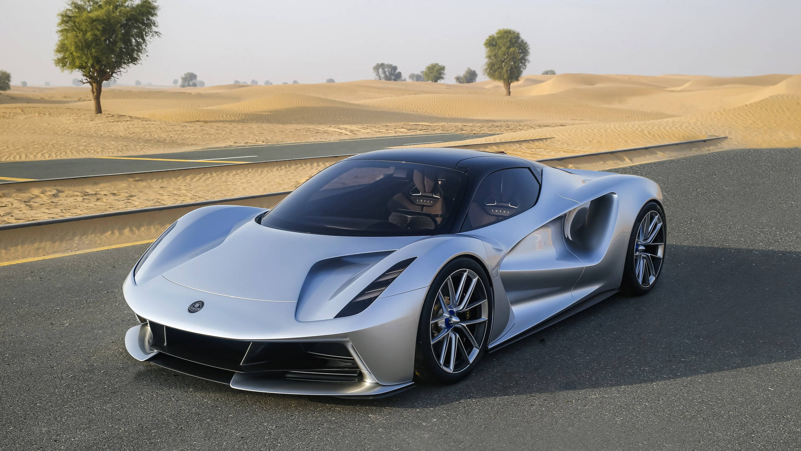 Sports Car 桌布 壁紙 — 一輛時尚的銀色跑車在沙漠公路上行駛，兩旁是金色的沙丘和稀疏的樹木，天空朦朧。