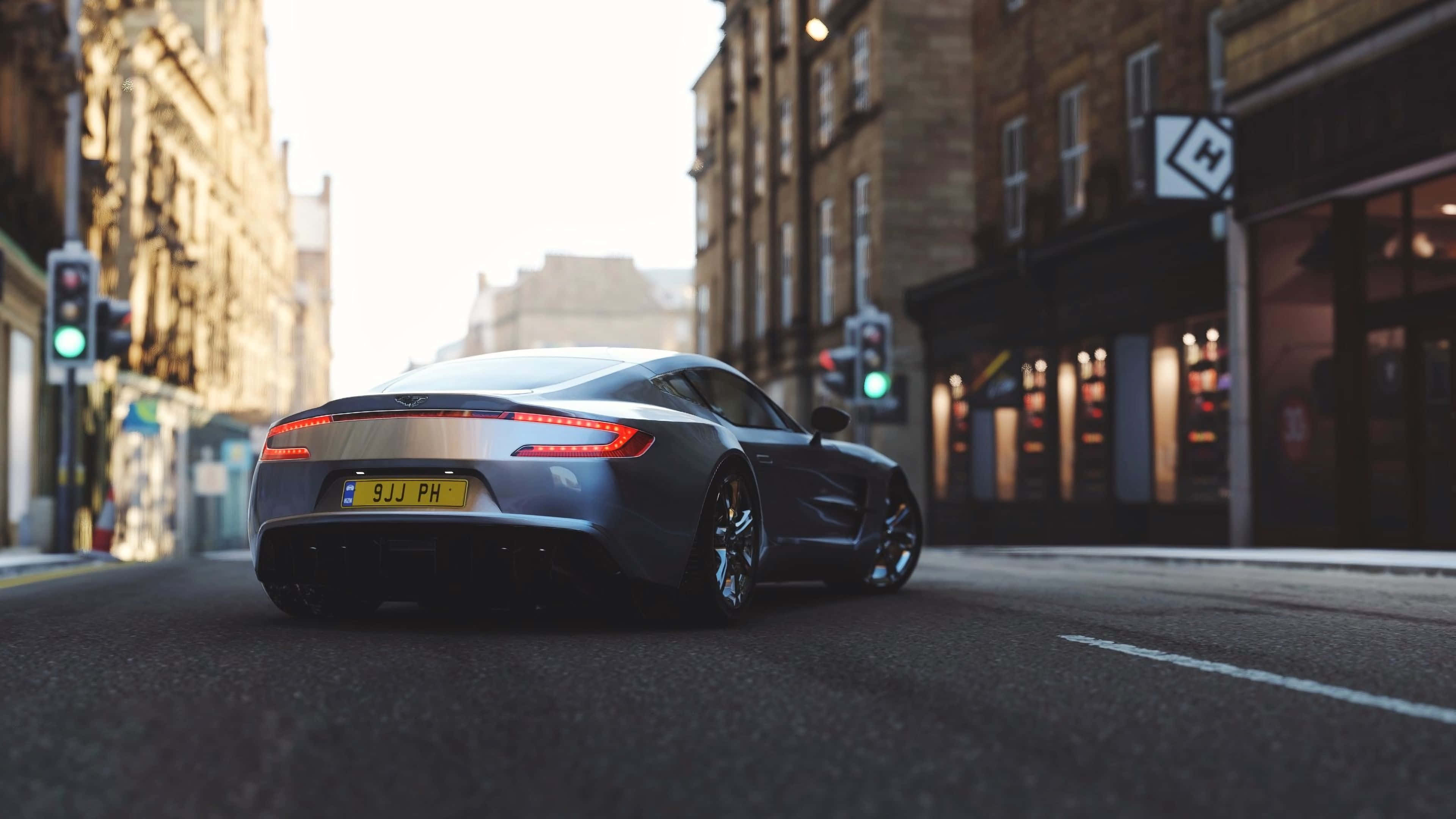 Aston Martin One-77 桌布 壁紙 — 一輛時尚的銀色跑車在陽光普照的市區街道上滑行，兩旁是歷史悠久的磚砌建築和閃閃發光的商店櫥窗。