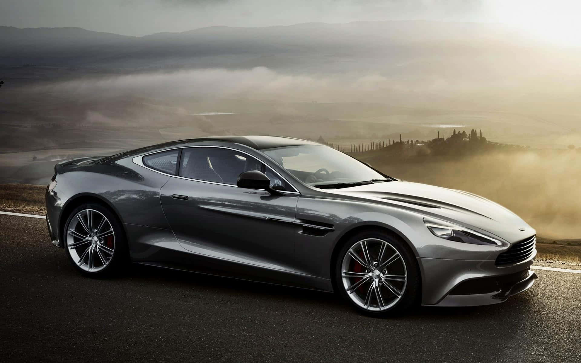 Aston Martin V12 Vantage 桌布 壁紙 — 一輛時尚的銀色跑車在黃金時段沿著蜿蜒的道路穿過霧氣瀰漫的山丘。