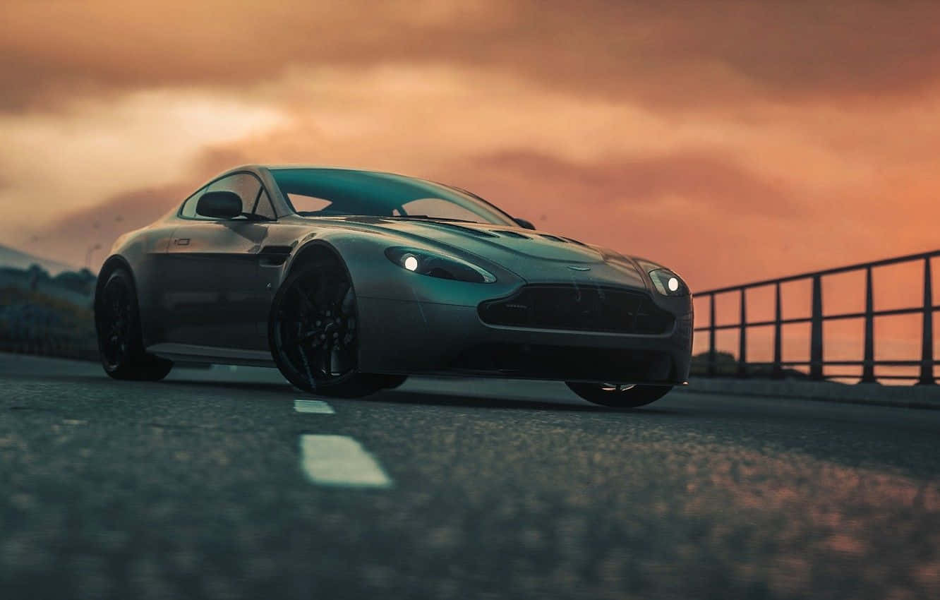 Aston Martin V12 Vantage 桌布 壁紙 — 一輛時尚的銀色跑車沿著海岸道路行駛，在火紅的夕陽天空下，伴隨著戲劇性的雲彩。
