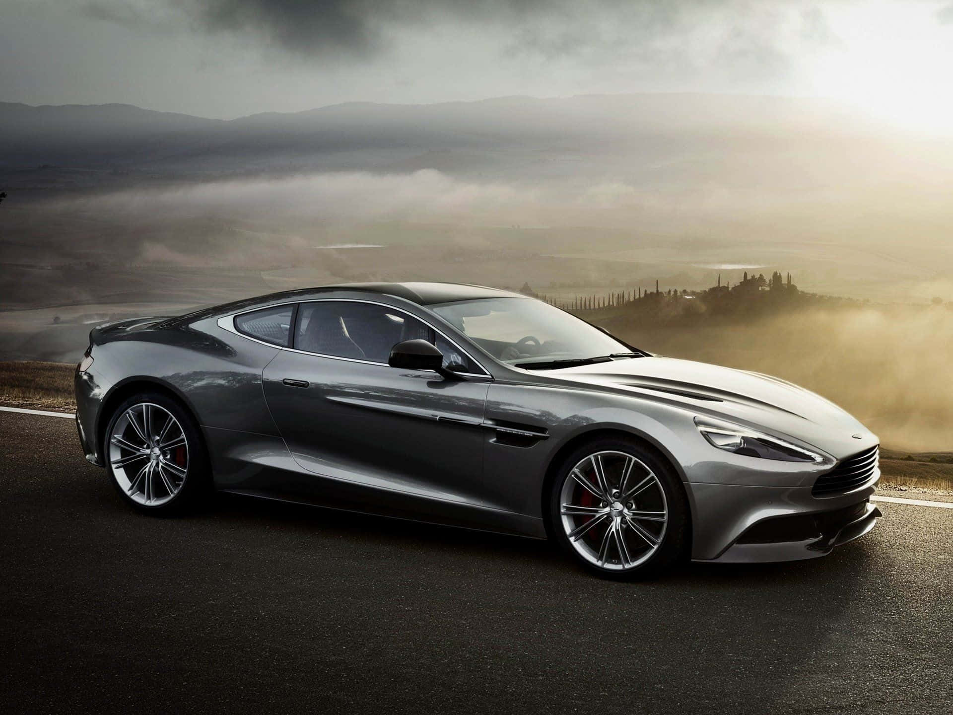 Aston Martin Vanquish 桌布 壁紙 — 一輛時尚的銀色跑車沿著蜿蜒的山路行駛，金色的日出光芒灑落在下方霧氣瀰漫的山谷中。