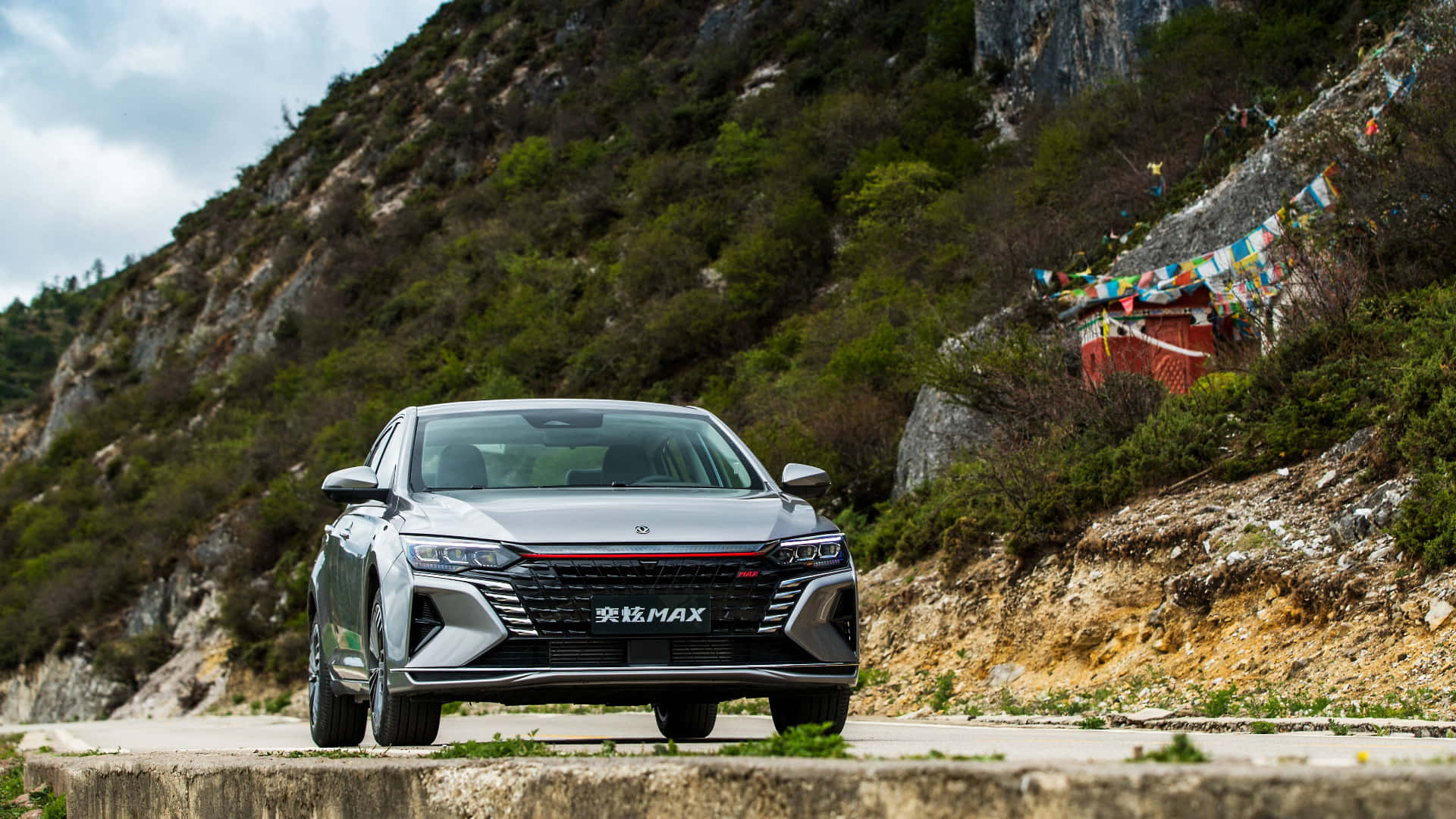 Dongfeng 桌布 壁紙 — 一輛時尚的銀色轎車停在路邊，背景是山巒，附近有色彩繽紛的經幡飄揚。