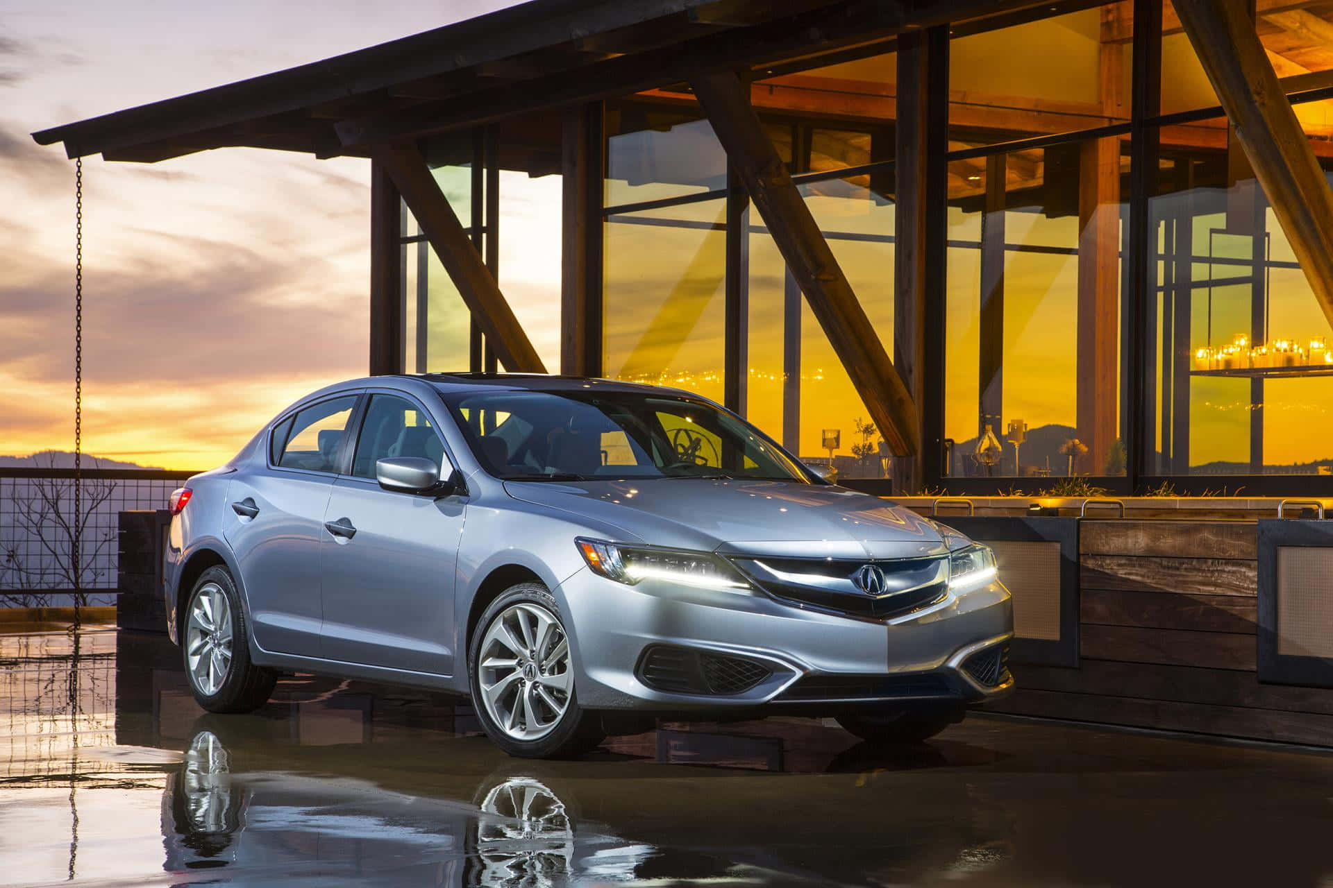 Acura Ilx 桌布 壁紙 — 一輛時尚的銀色轎車在日落時分的濕滑路面上閃閃發光，反射出現代玻璃建築溫暖的金色光芒。