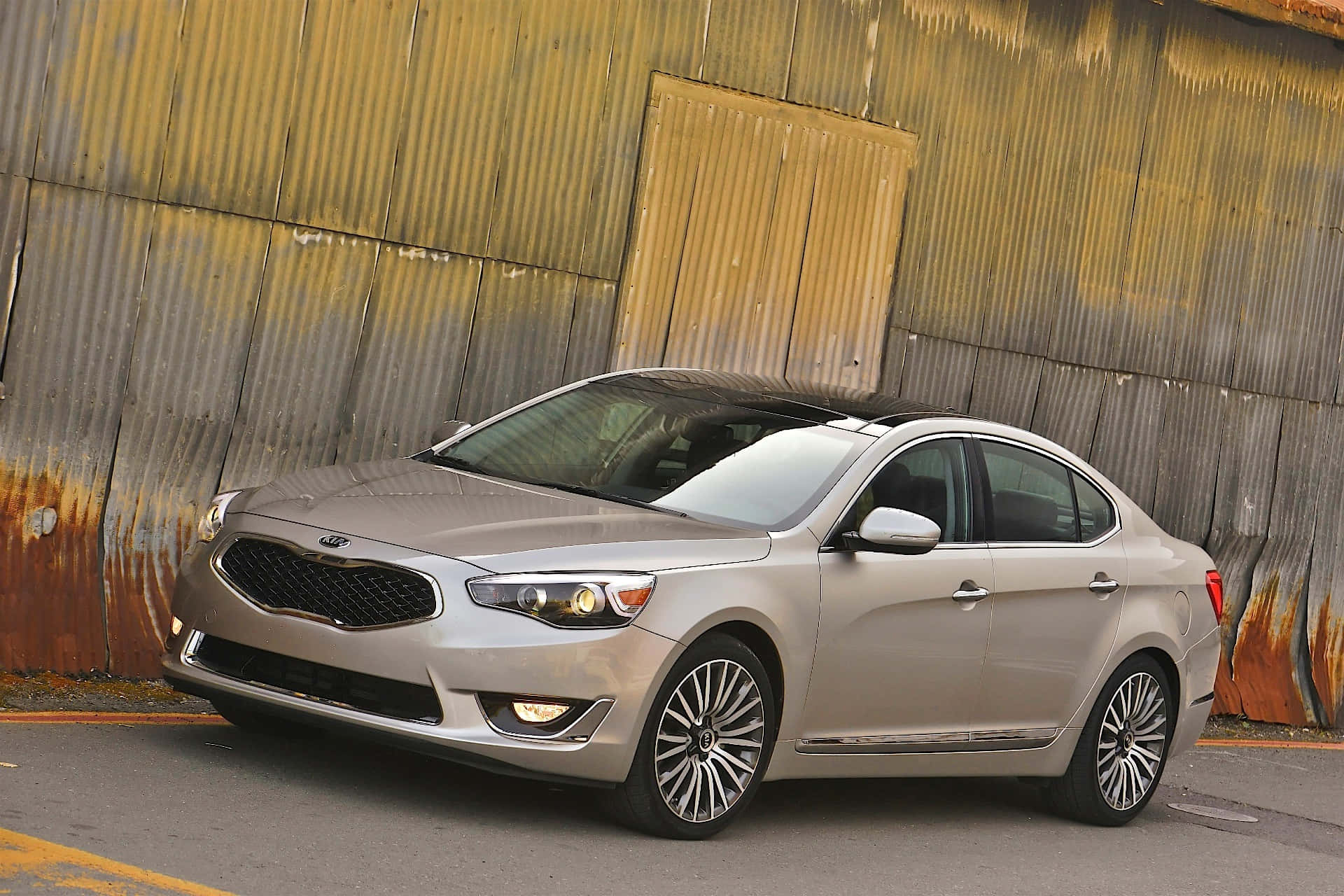 Kia Cadenza 桌布 壁紙 — 一輛時尚的銀色轎車在明亮的日光下停泊在風化生鏽的波紋金屬牆旁。