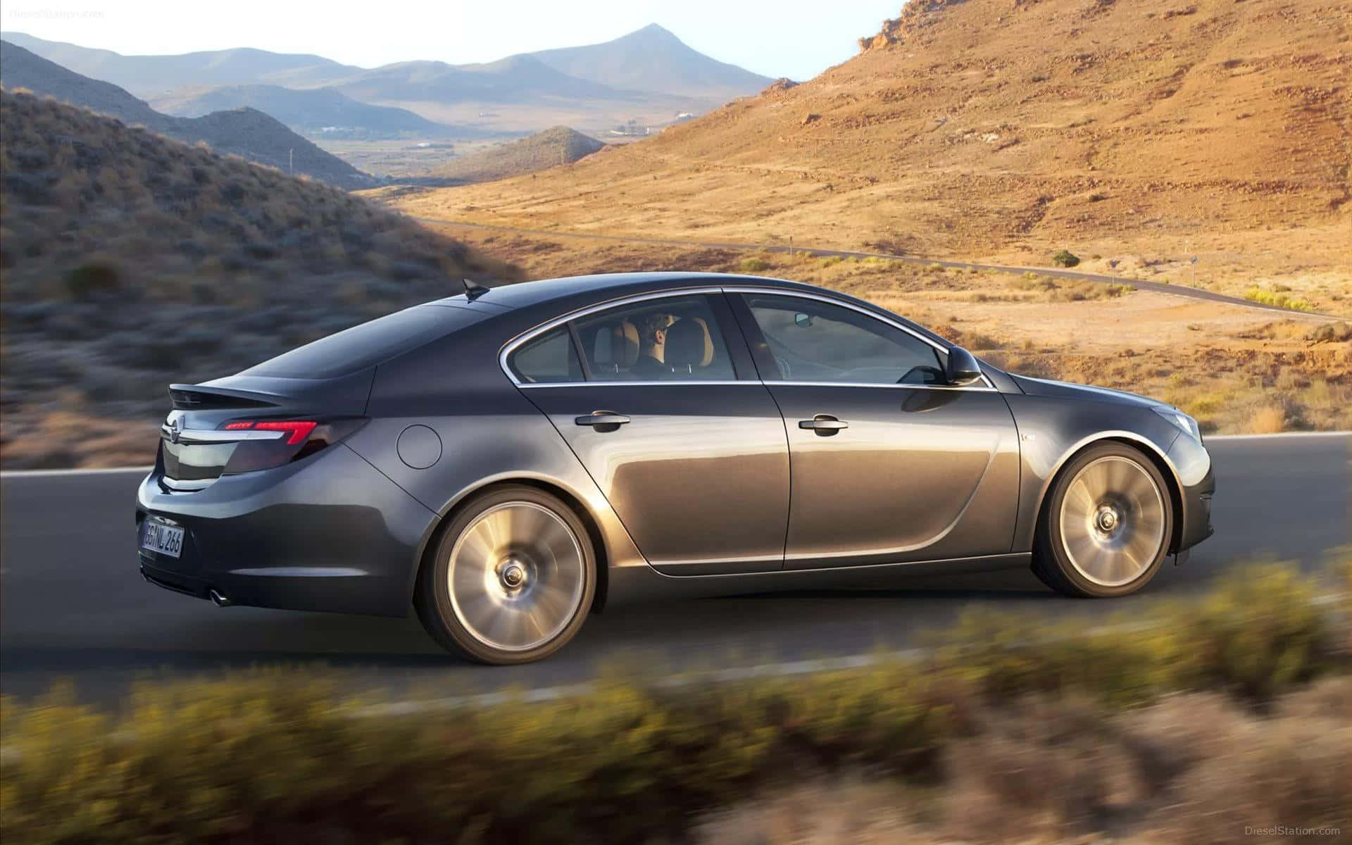 Opel Insignia 桌布 壁紙 — 一輛時尚的銀色轎車在蜿蜒的沙漠公路上疾馳，背景是崎嶇的山脈，沐浴在金色的午後陽光中。