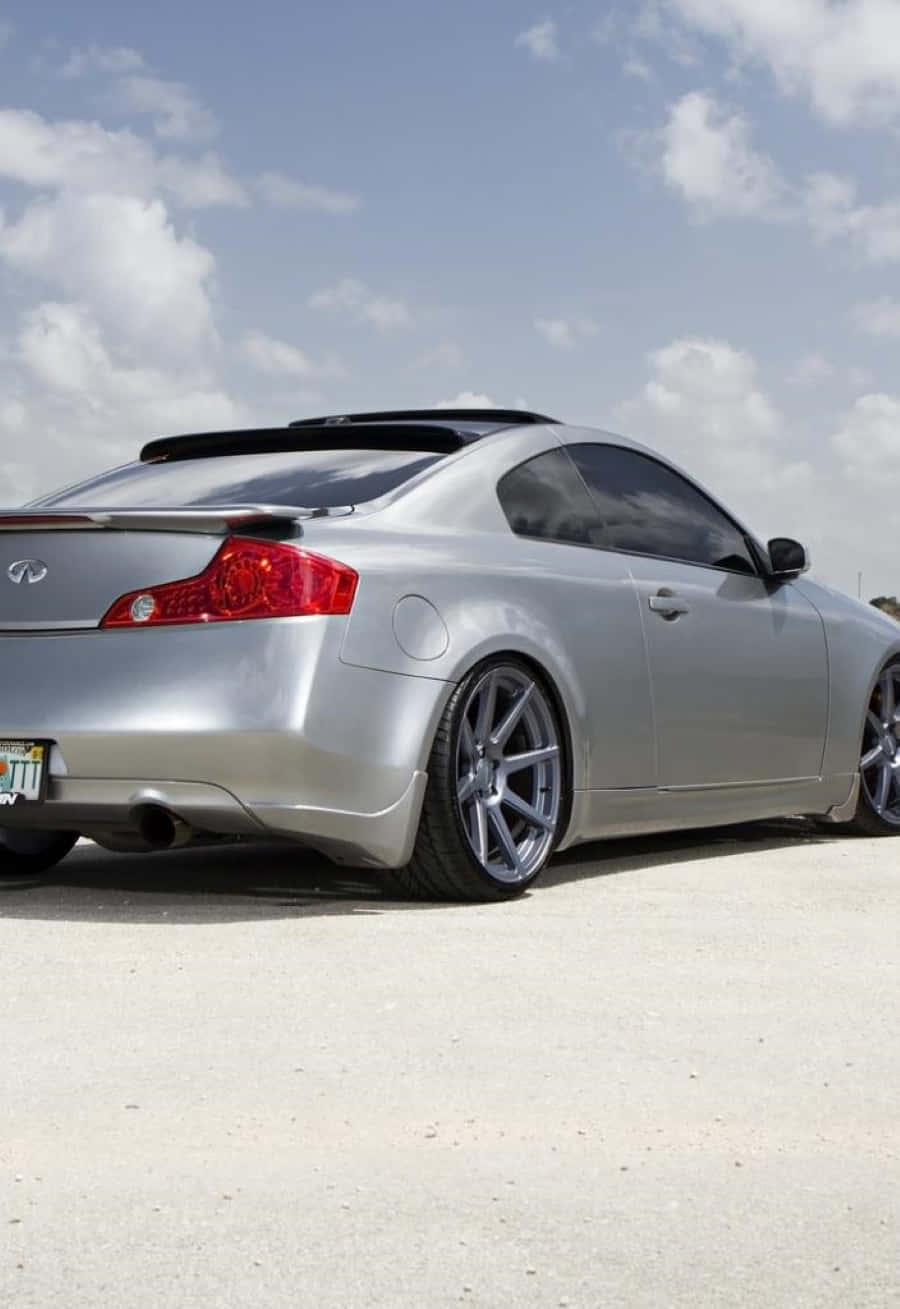 Infiniti G35 桌布 壁紙 — 一輛時尚的銀色運動轎跑車，配備定制輪圈，停放在混凝土場地上，天空部分多雲。