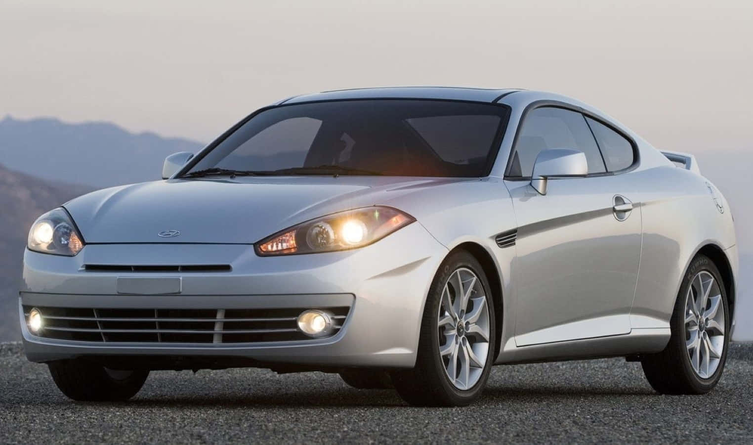 Hyundai Tiburon 桌布 壁紙 — 一輛時尚的銀色運動轎跑車停在碎石路上，車頭燈亮起，背景是黃昏時分朦朧的山巒。