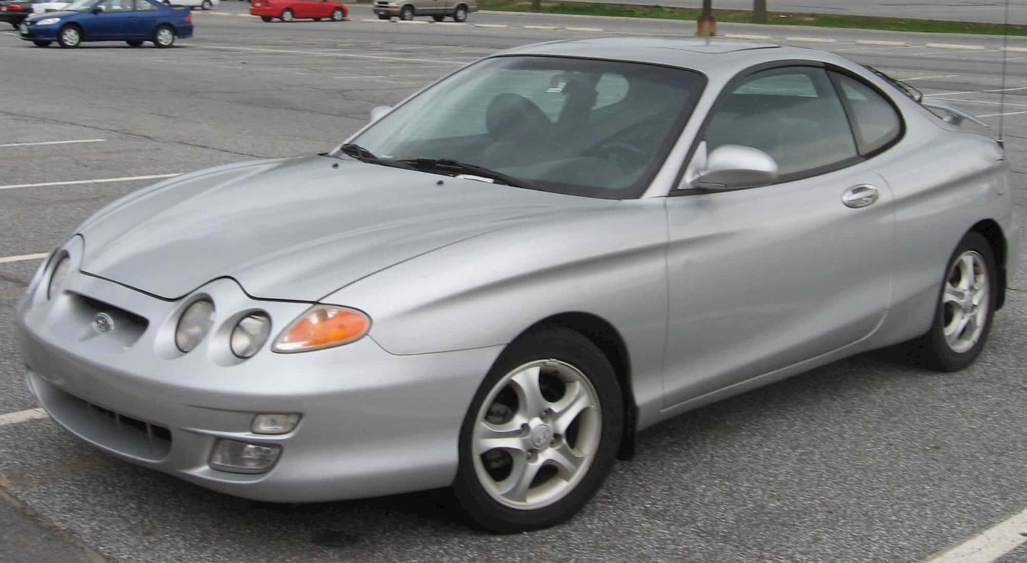 Hyundai Tiburon 桌布 壁紙 — 一輛時尚的銀色雙門跑車停放在碎石停車場，背景可見其他車輛。