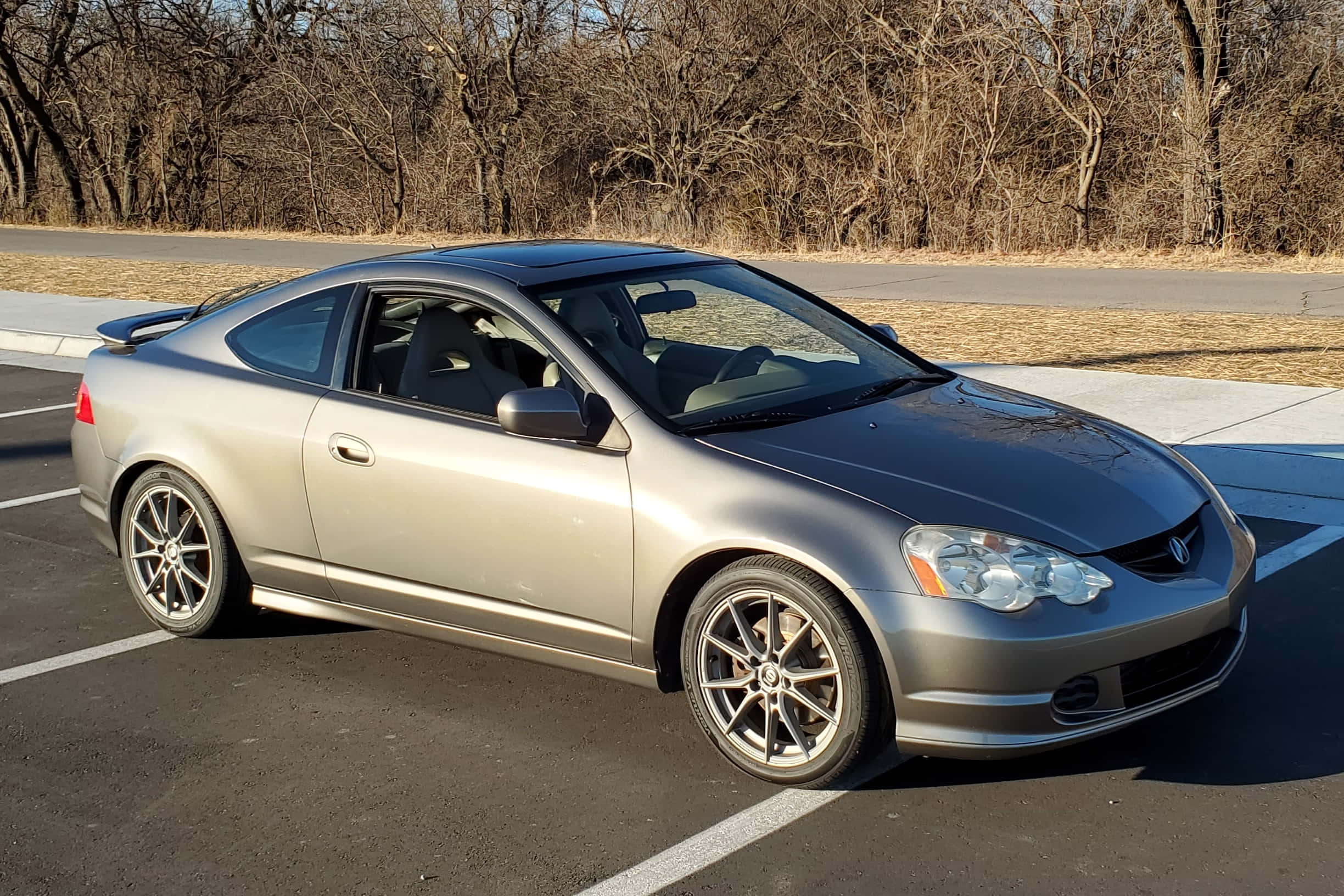 Acura Rsx 桌布 壁紙 — 一輛時尚的銀色雙門轎跑車停放在陽光明媚的停車場，背景是光禿禿的樹木。