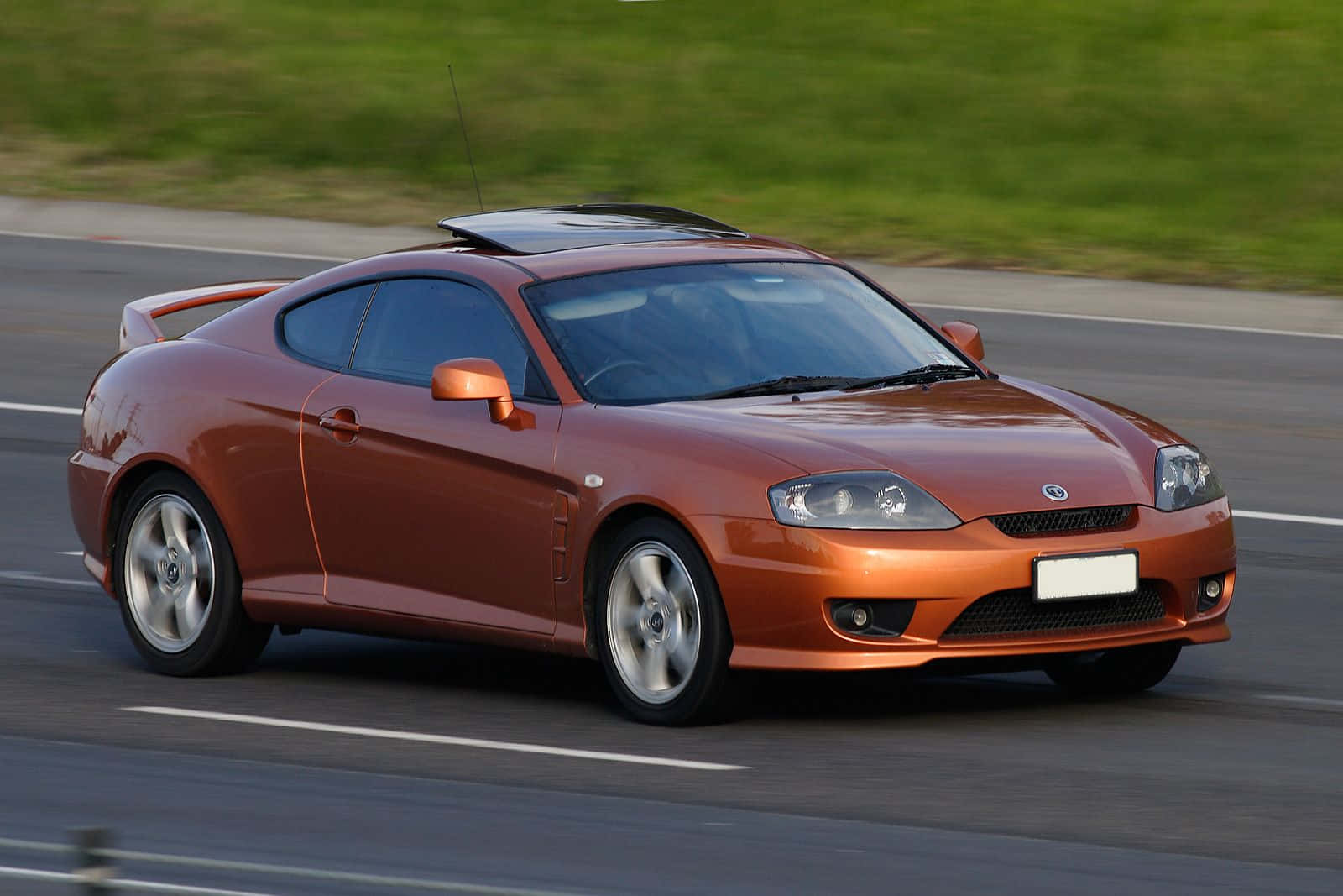 Hyundai Tiburon 桌布 壁紙 — 一輛時尚的銅色跑車在鋪砌的道路上飛馳，背景是模糊的綠色草地。