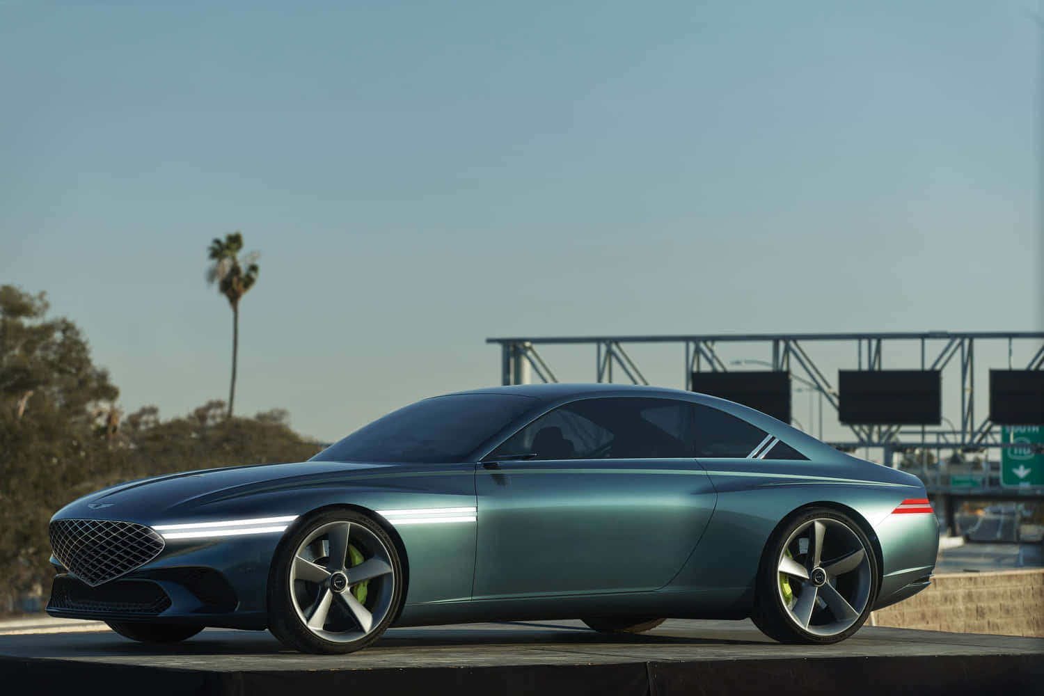 Genesis X Concept 桌布 壁紙 — 一輛時尚的青色概念車帶有發光飾件，在晴朗藍天下停在高速公路天橋上。