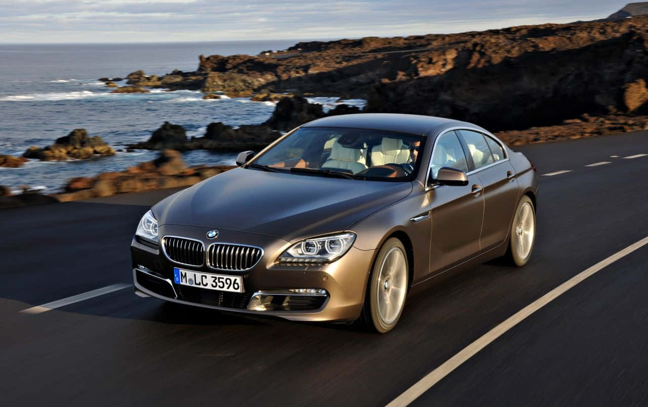 Bmw 6 Series 桌布 壁紙 — 一輛時尚的青銅色BMW 6系轎跑車沿著海岸公路行駛，背景是崎嶇的懸崖和拍岸的浪花。