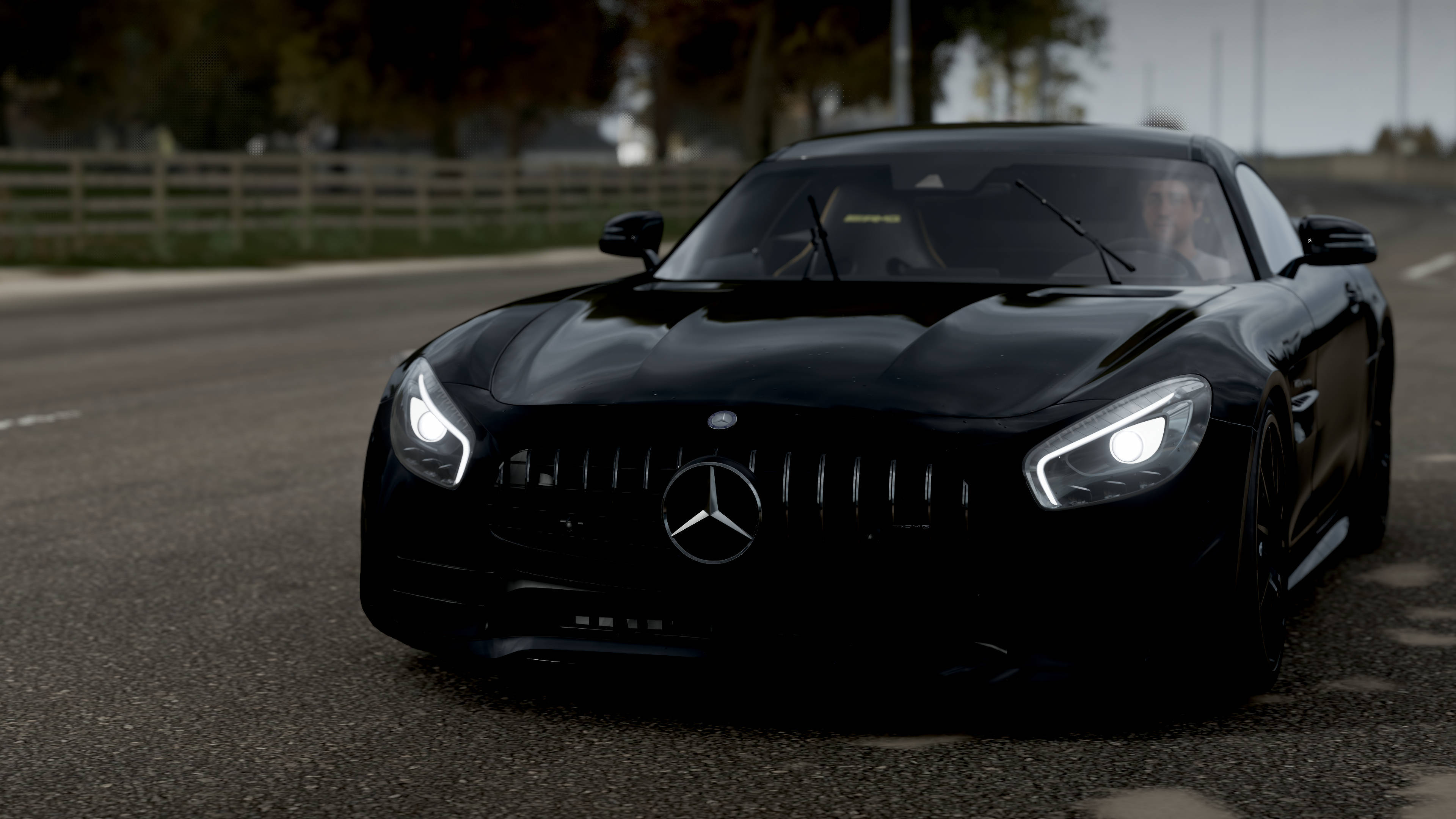 遊戲 桌布 壁紙 — 一輛時尚的黑色 Mercedes-AMG GT 跑車，帶有發光的車頭燈，停在木柵欄附近的瀝青路上。