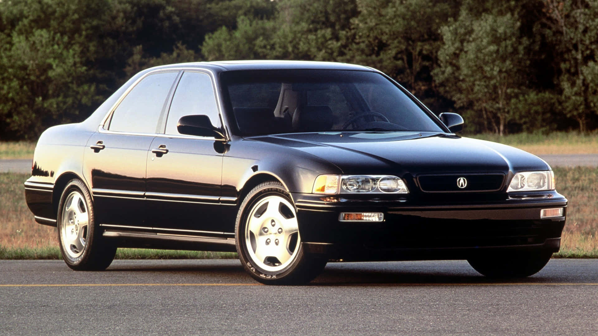 Acura Legend 桌布 壁紙 — 一輛時尚的黑色Acura轎車在金色陽光下閃閃發光，停在有樹木背景的瀝青路上。