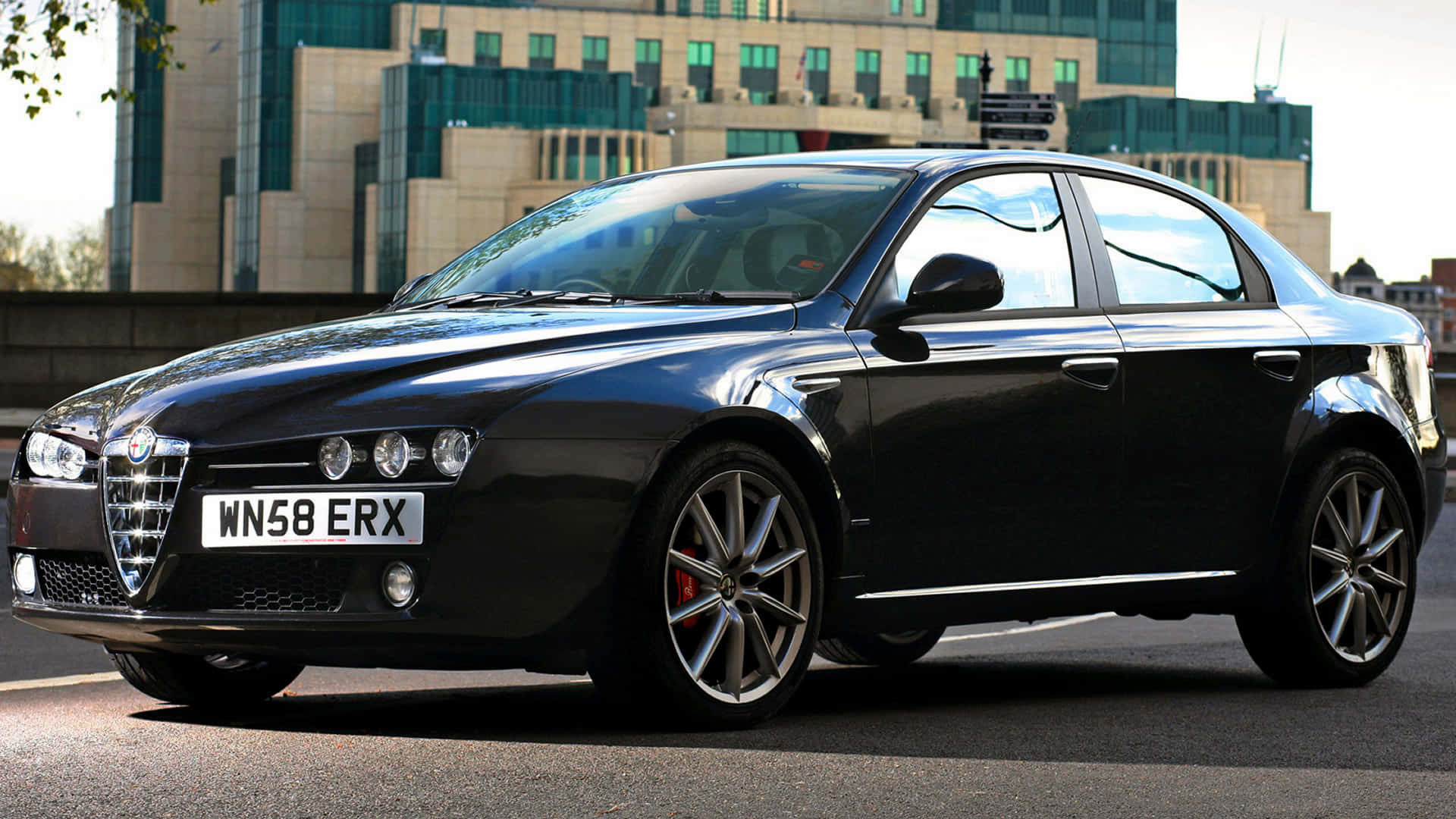 Alfa Romeo 159 桌布 壁紙 — 一輛時尚的黑色Alfa Romeo房車在日光下閃耀，背景是現代建築的城市街道。