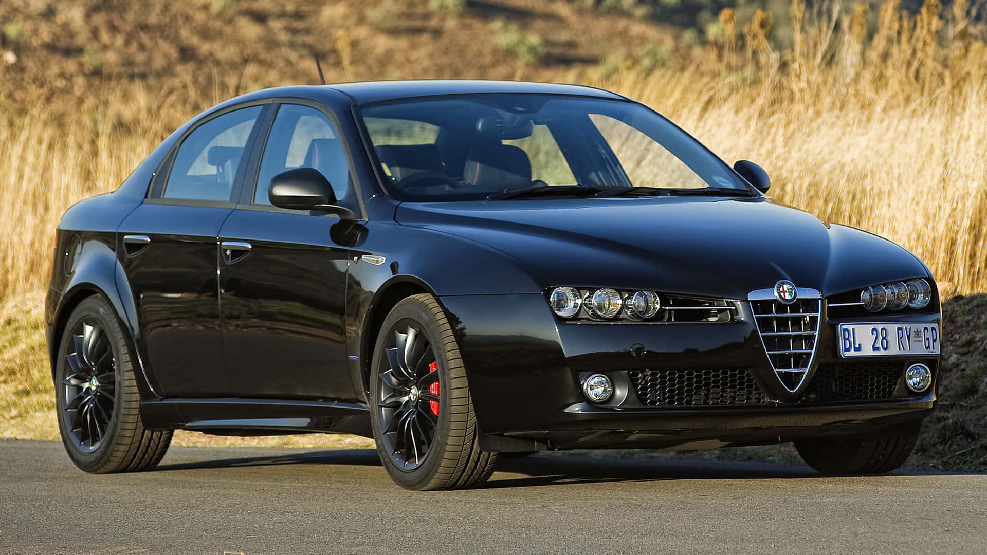 Alfa Romeo 159 桌布 壁紙 — 一輛時尚的黑色Alfa Romeo轎車，帶紅色制動卡鉗，停在鋪砌的道路旁，旁邊是金色的草。