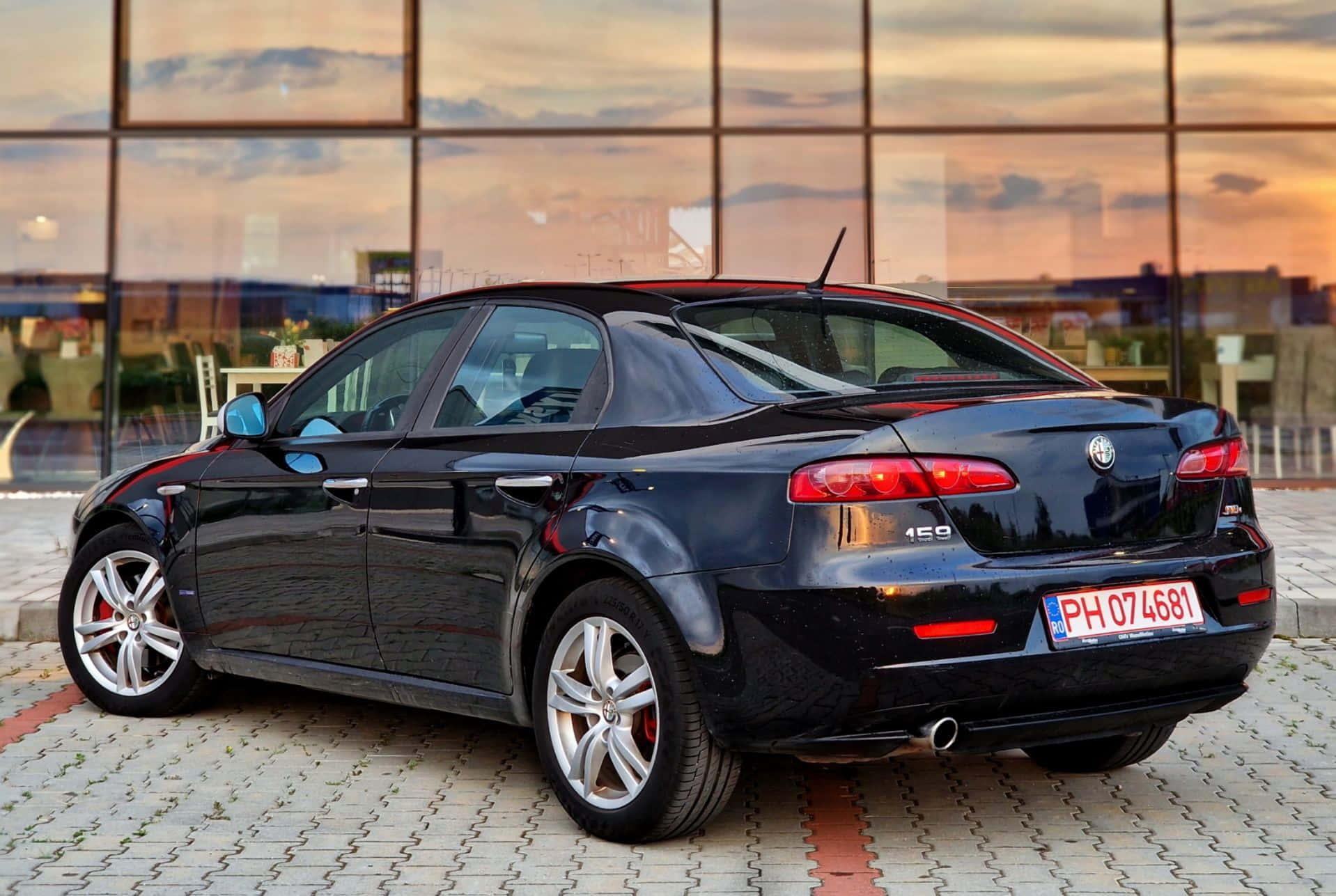 Alfa Romeo 159 桌布 壁紙 — 一輛時尚的黑色Alfa Romeo 156轎車停泊在鋪砌的廣場上，夕陽西下，其光澤表面反射著溫暖的橙色天空。