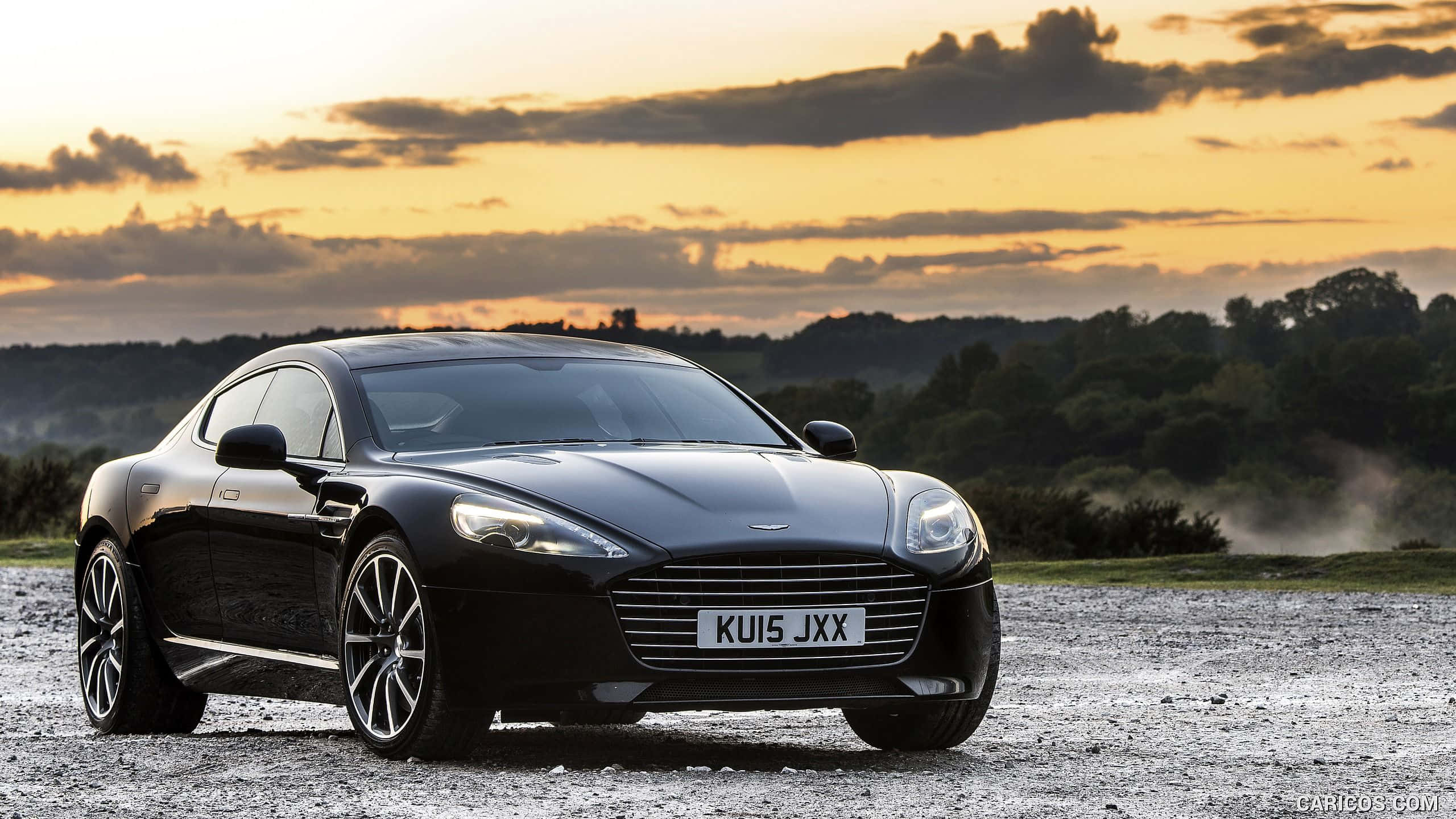 Aston Martin Rapide S 桌布 壁紙 — 一輛時尚的黑色Aston Martin DB9停在碎石上，夕陽西下，金色雲彩和遠處的樹木。