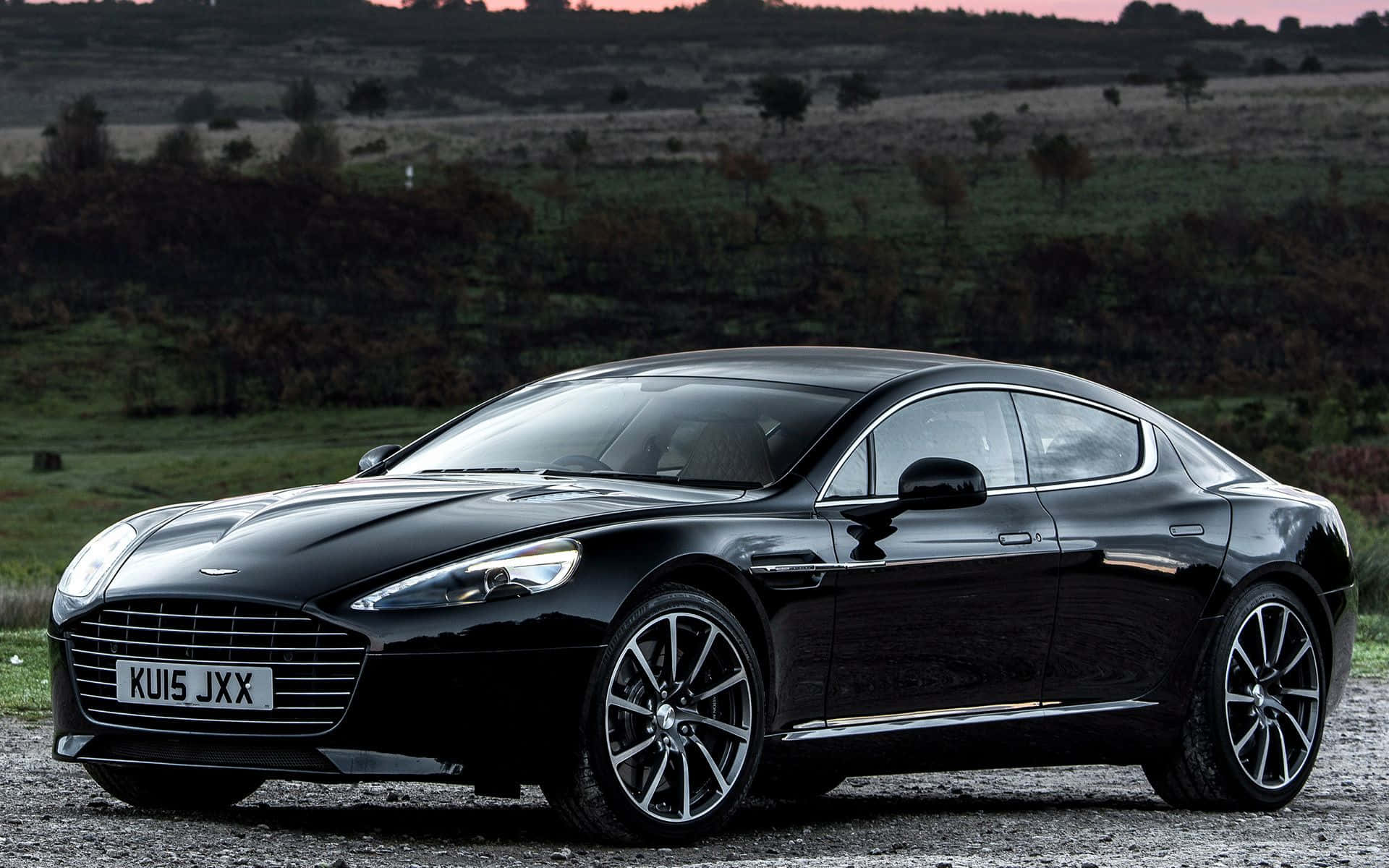 Aston Martin Rapide S 桌布 壁紙 — 一輛時尚的黑色Aston Martin跑車在暮色中閃耀，停在碎石路上，背景是起伏的綠色山丘。