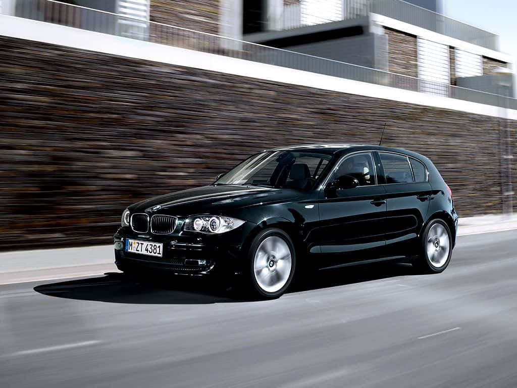 Bmw 1 Series 桌布 壁紙 — 一輛時尚的黑色BMW 1系掀背車在現代城市道路上疾馳，石牆和行人路上呈現動態模糊效果。
