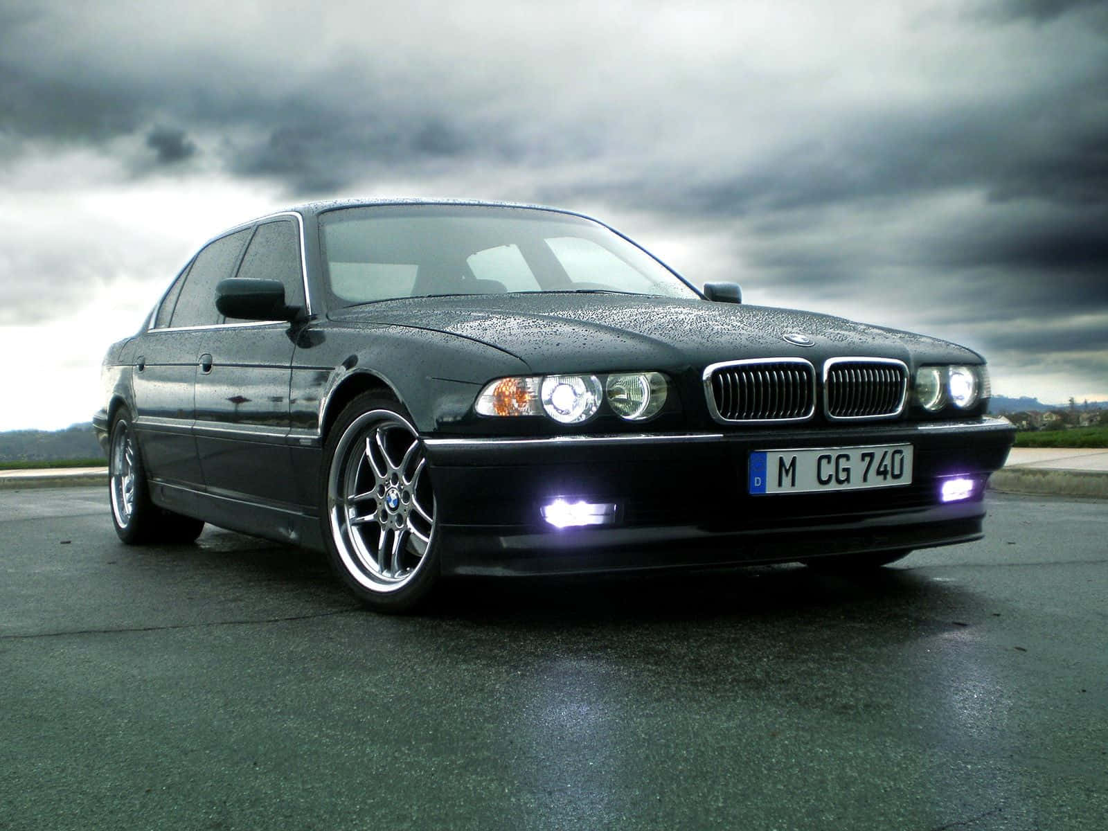 Bmw 7 Series 桌布 壁紙 — 一輛時尚的黑色BMW 7系轎車在陰天中閃耀，引擎蓋上有雨滴，車頭燈亮起。