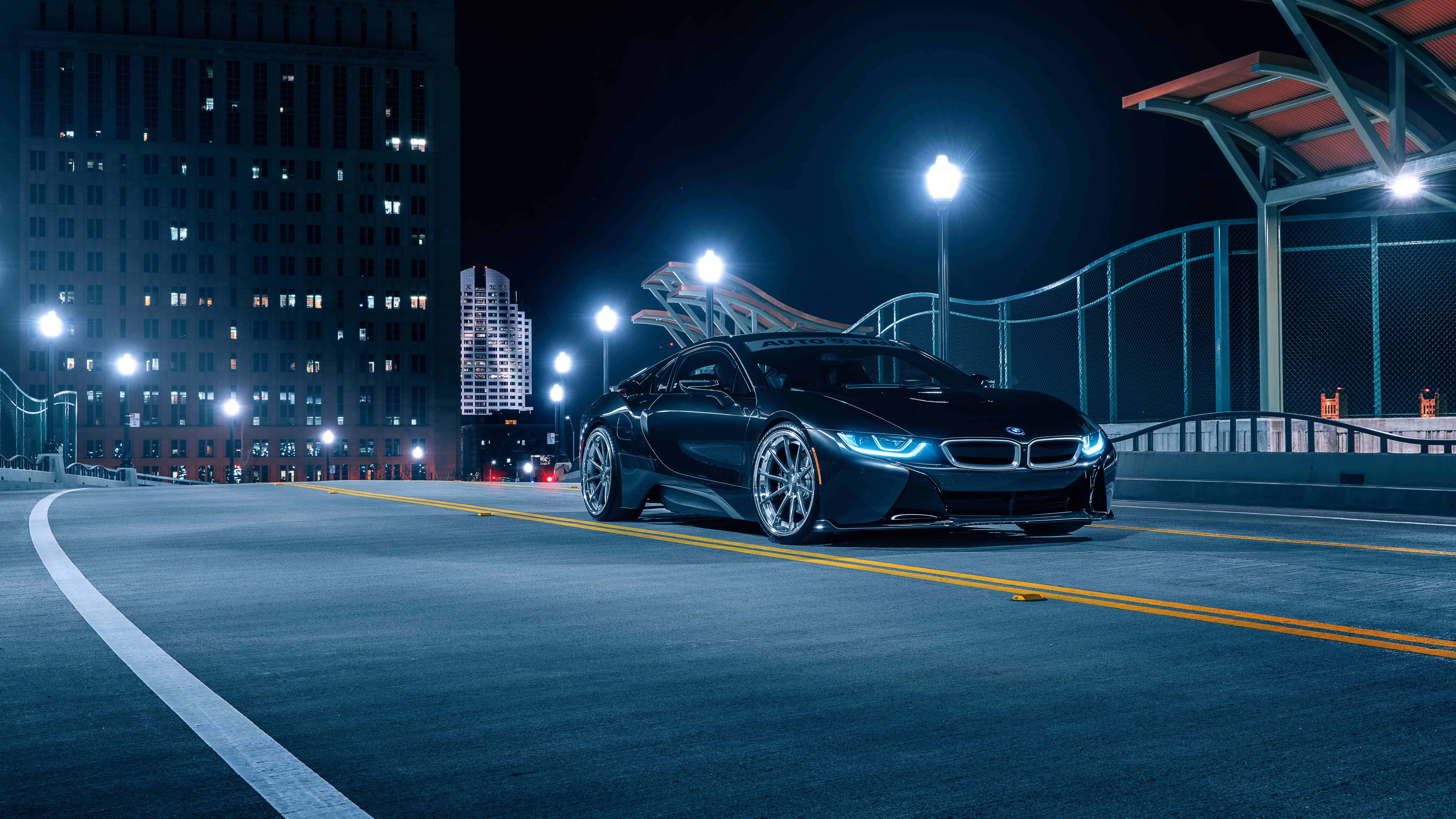 8K 桌布 壁紙 — 一輛時尚的黑色BMW i8跑車在夜晚彎曲的城市道路上，在霓虹燈下閃耀，背景是高樓大廈。