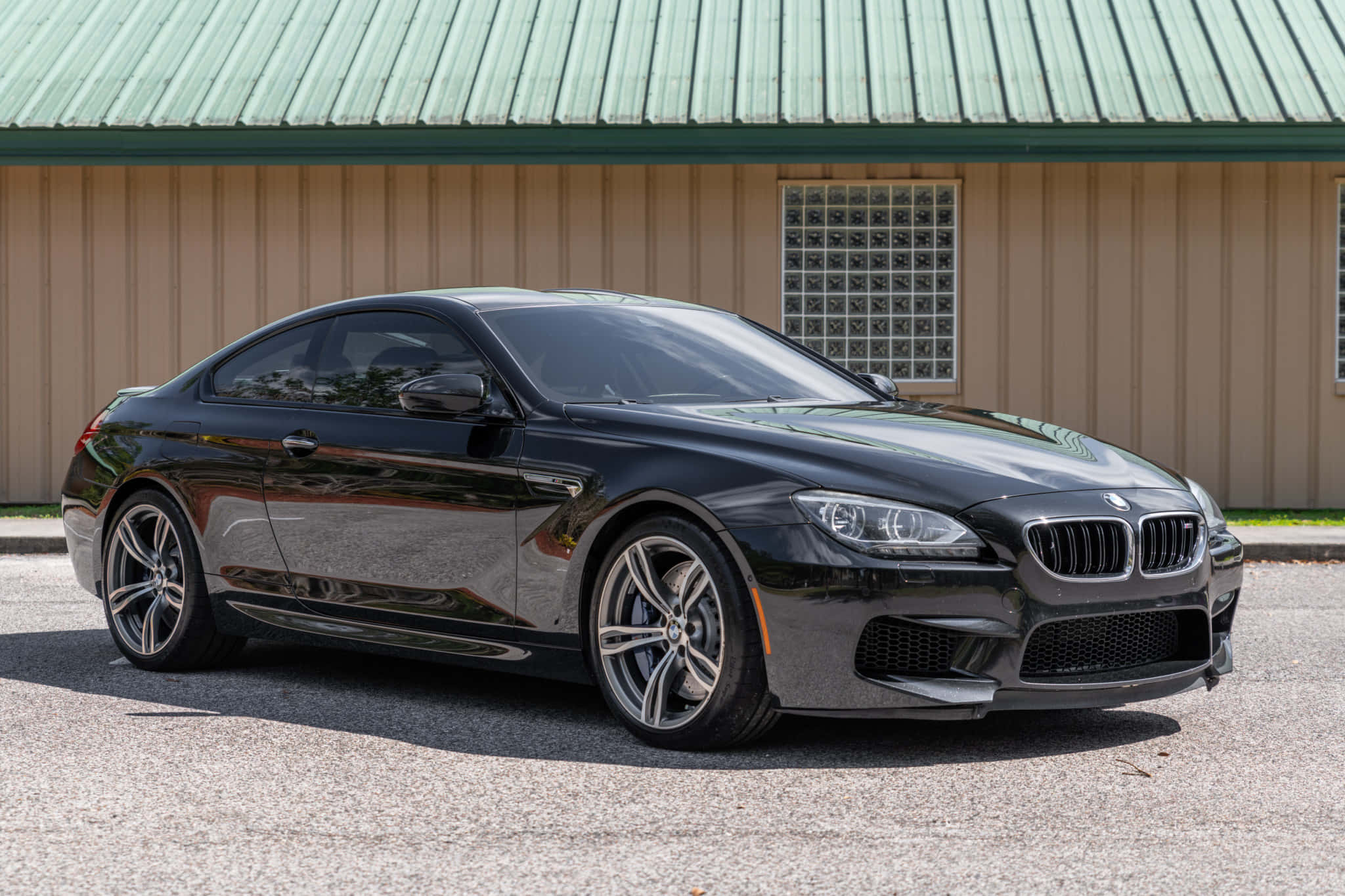 Bmw M6 桌布 壁紙 — 一輛時尚的黑色BMW M6轎跑車在明亮的陽光下閃閃發光，停在瀝青路面上，前方是米色建築和綠色金屬屋頂。