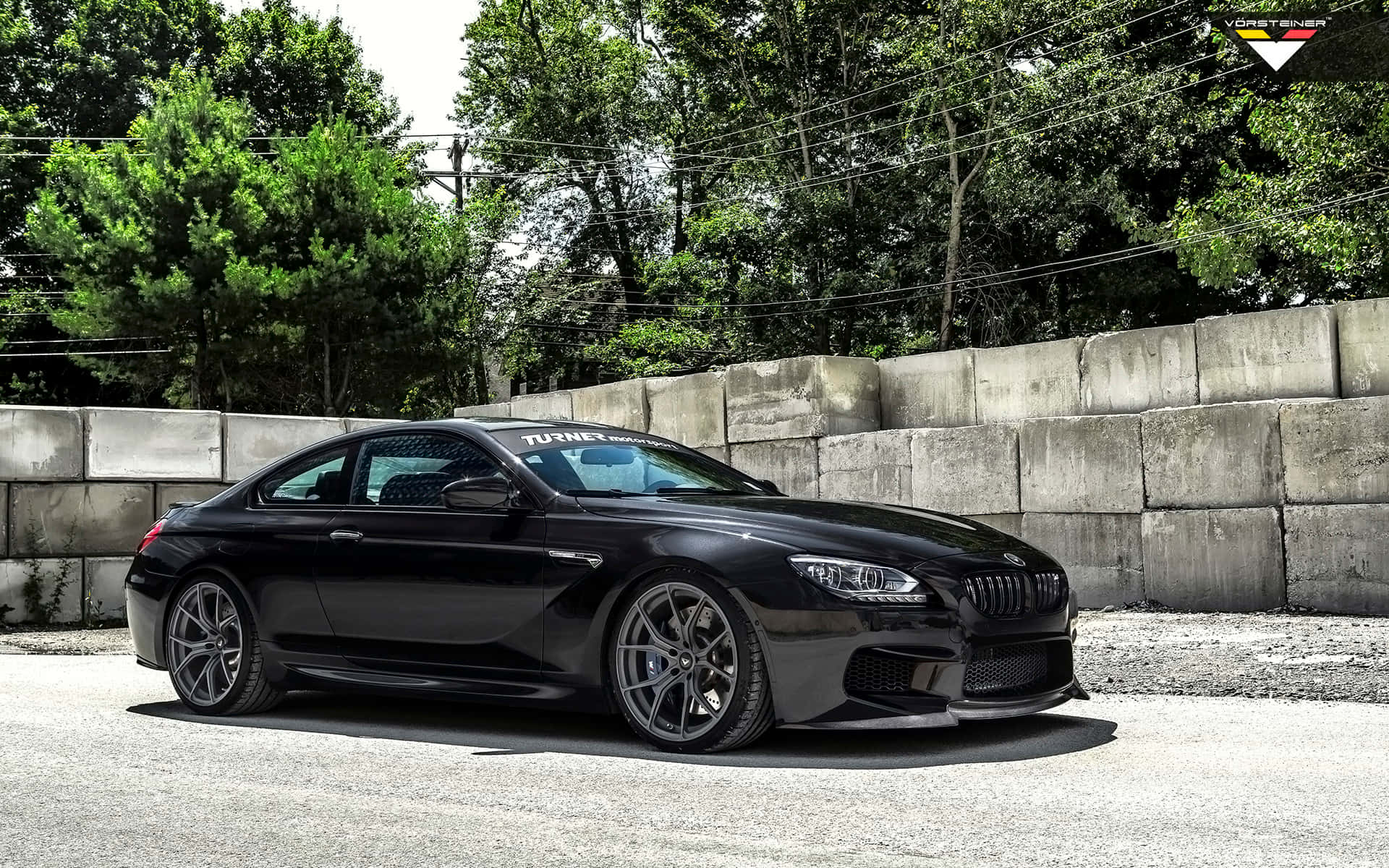 Bmw M6 桌布 壁紙 — 一輛時尚的黑色BMW M6雙門跑車停在碎石上，旁邊是混凝土牆，背景有茂密的綠樹。