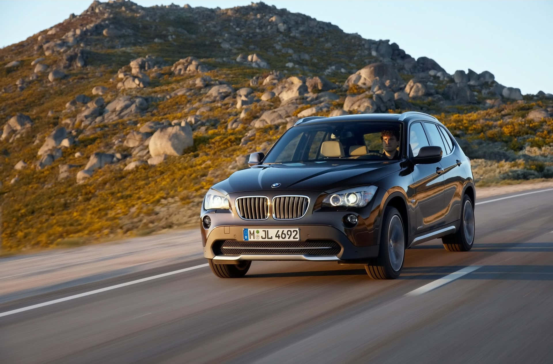 Bmw I8 桌布 壁紙 — 一輛時尚的黑色BMW X1 SUV在蜿蜒的道路上行駛，穿過沐浴在金色日落光線中的岩石山坡。