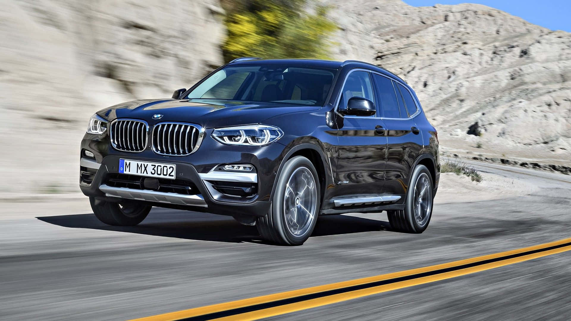 Bmw X3 桌布 壁紙 — 一輛時尚的黑色BMW X3 SUV在蜿蜒的沙漠公路上疾馳，背景是模糊的岩石山丘。