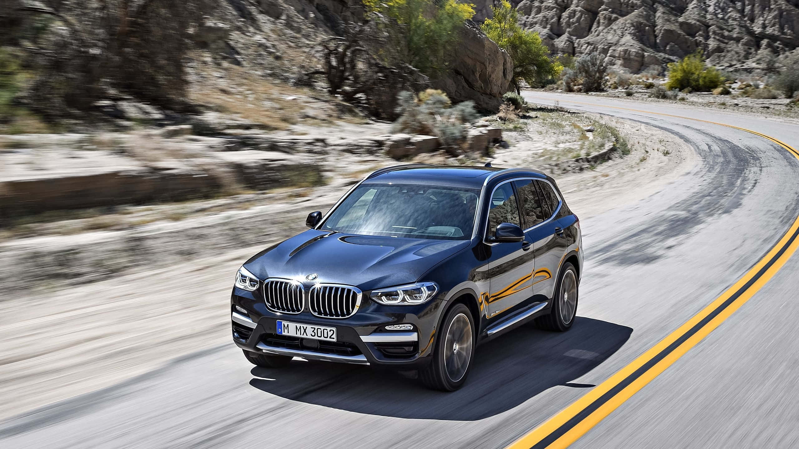 Bmw X3 桌布 壁紙 — 一輛時尚的黑色BMW X3 SUV在蜿蜒的沙漠公路上疾馳，沿途有岩石峭壁和稀疏的綠植，呈現動態模糊效果。