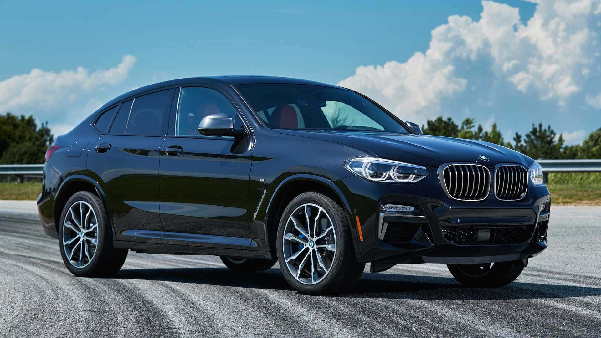 Bmw X4 桌布 壁紙 — 一輛時尚的黑色BMW X4 SUV在明亮的陽光下，在開闊的道路上閃閃發光，背景是蓬鬆的白雲和綠樹。