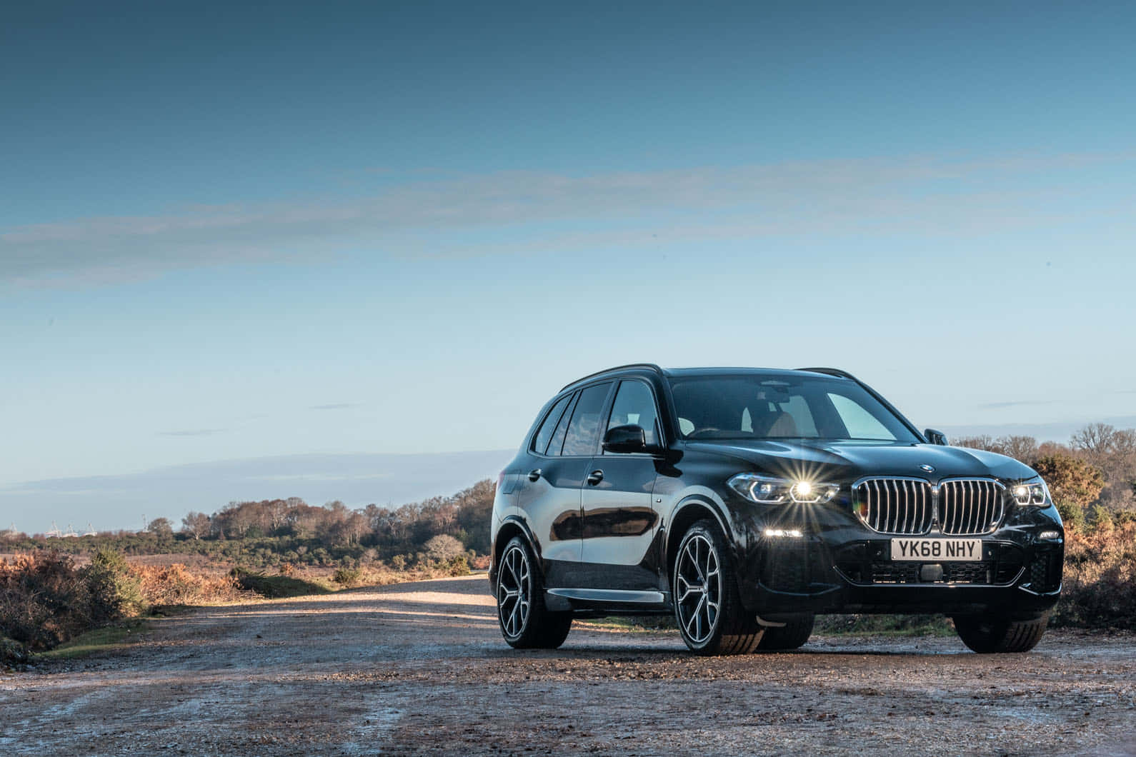 Bmw X5 桌布 壁紙 — 一輛時尚的黑色BMW X5 SUV在晴朗的藍天下，在穿過秋日鄉村的碎石路上閃閃發光。