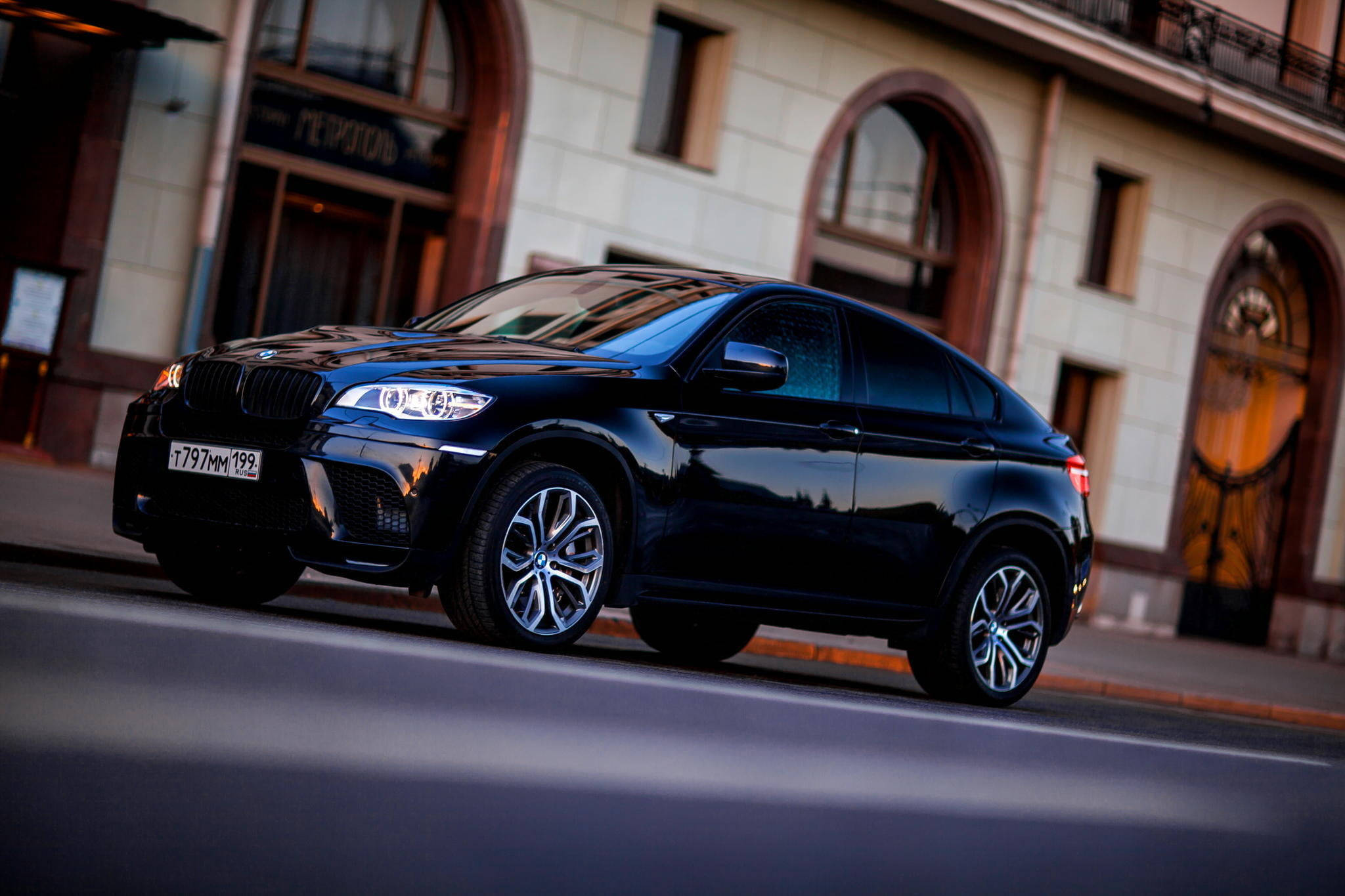 BMW X6 M SUV 壁紙 — 一輛時尚的黑色BMW X6運動型轎跑車在黃昏時分沿著城市街道滑行，溫暖的建築燈光反射在其光滑的表面上。