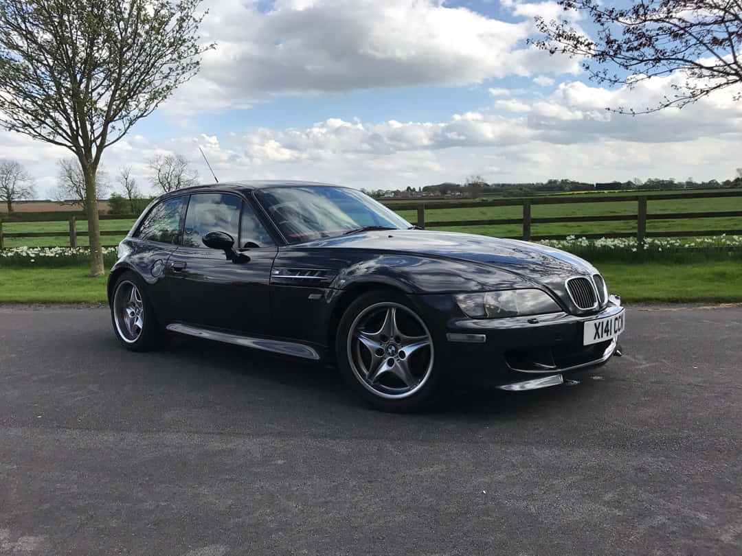 Bmw Z3 桌布 壁紙 — 一輛時尚的黑色BMW Z3跑車停泊在鋪砌的車道上，背景是綠色的田野和盛開的白花，天空部分多雲。