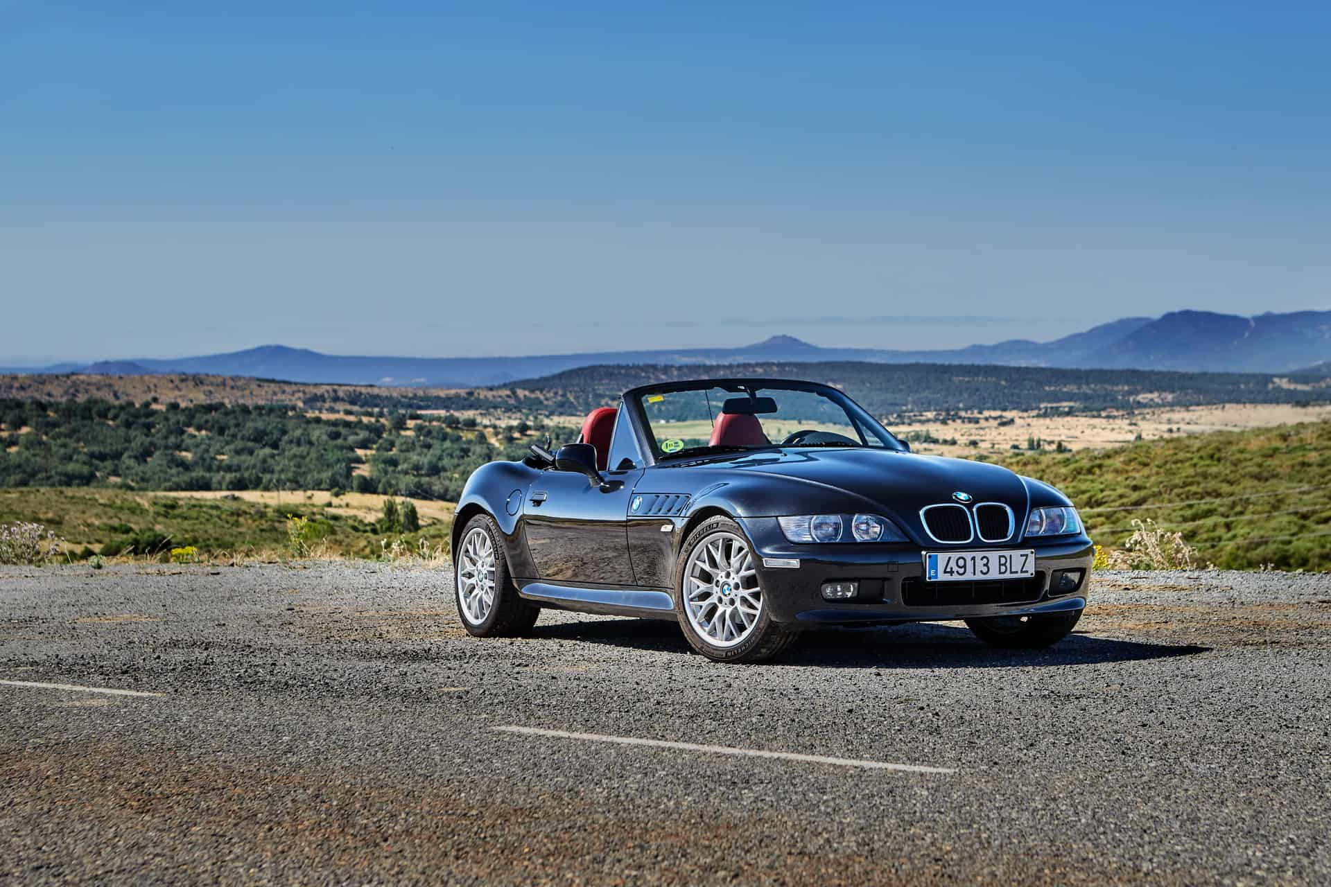 Bmw Z3 桌布 壁紙 — 一輛時尚的黑色BMW Z3開篷跑車，配紅色座椅，停泊在碎石路上，俯瞰連綿起伏的山丘和遠處的山脈，天空清澈湛藍。